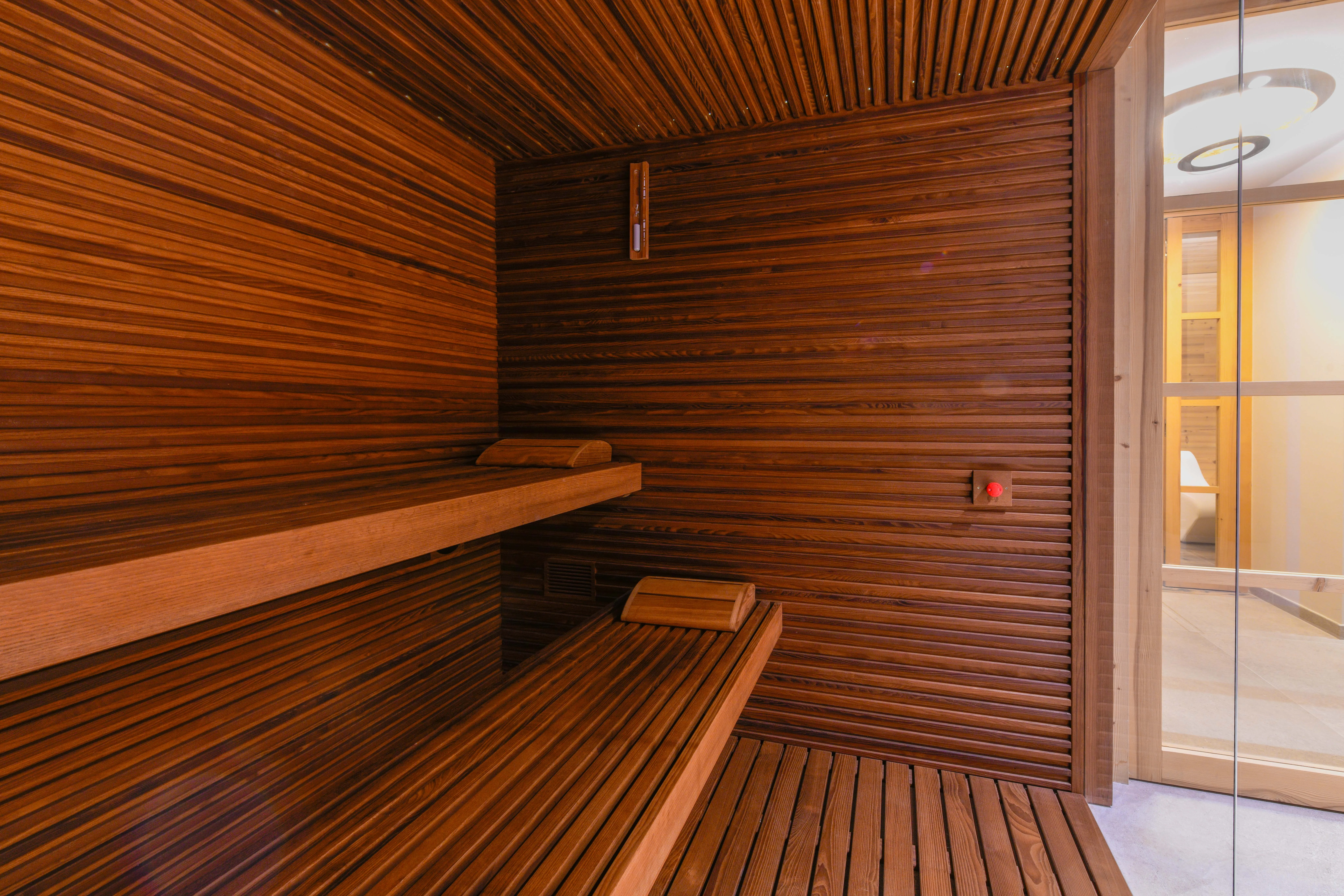 Holzverkleidete Sauna mit zwei Sitzbänken aus Holz auf unterschiedlichen Höhen. Die Tür zur Sauna ist aus Glas und zeigt einen hell erleuchteten Vorraum.