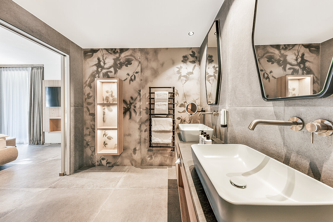 Modernes Badezimmer mit zwei rechteckigen Waschbecken auf einer Holzplatte, zwei Spiegeln, einem wandmontierten Wasserhahn und dekorativer Wandgestaltung. Im Hintergrund ein Handtuchhalter und stimmungsvolle Wandbeleuchtung mit floralen Motiven. Links öffnet sich der Raum zu einem Wohnbereich.