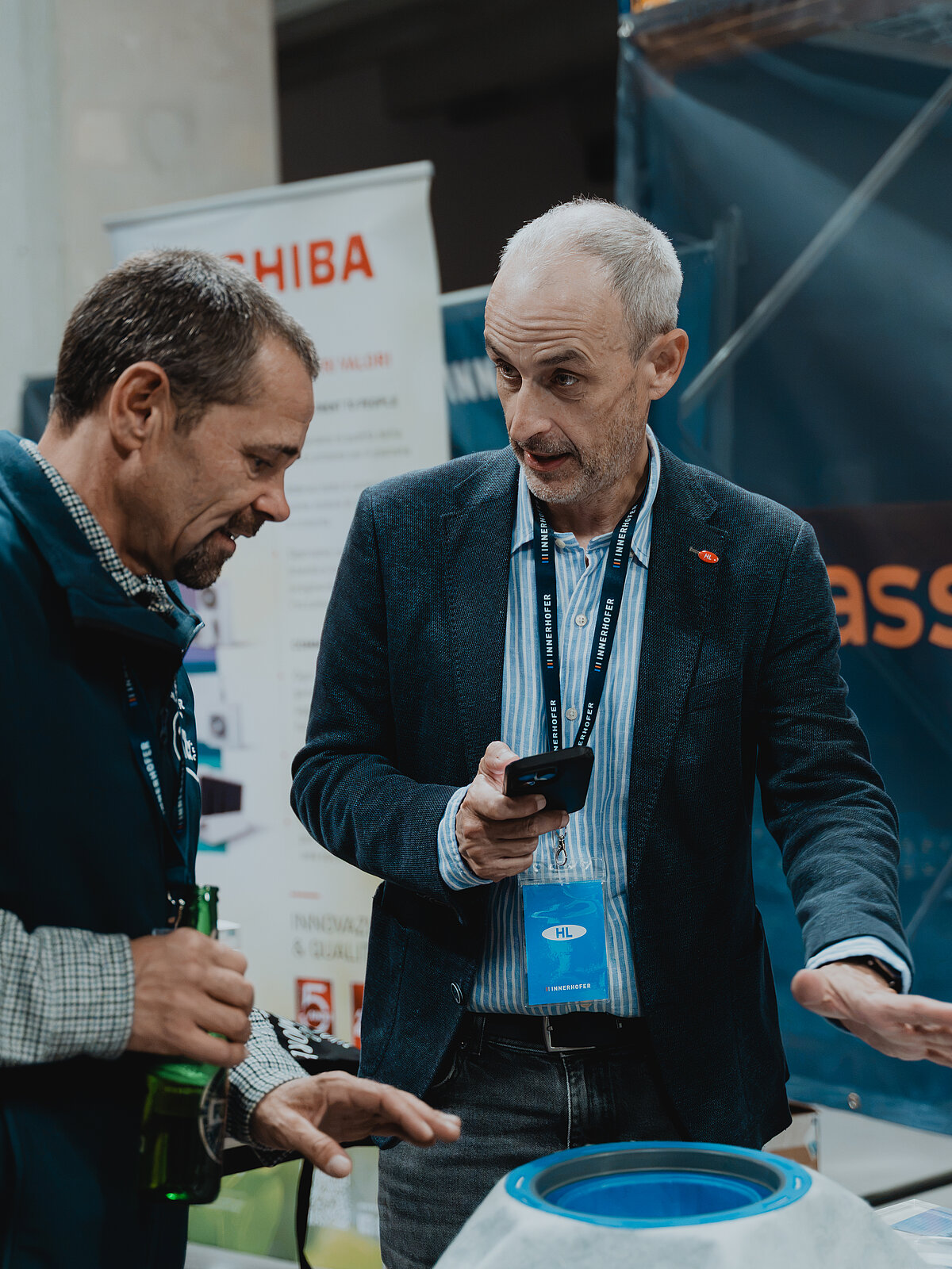 Zwei Männer stehen an einem Messestand und unterhalten sich. Einer hält ein Smartphone, während der andere eine Flasche in der Hand hat. Im Hintergrund sind Werbebanner zu sehen. Beide scheinen sich intensiv über ein ausgestelltes Produkt auszutauschen.