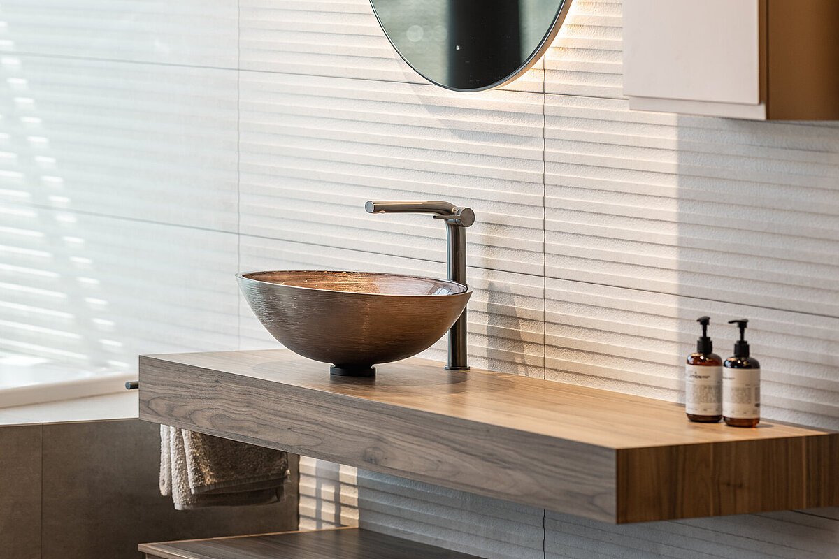 Modernes Badezimmer mit einem runden Spiegel über einem Holzwaschtisch. Eine runde Waschschüssel und ein Wasserhahn befinden sich auf dem Waschtisch. Zwei Seifenspender stehen daneben. Darunter sind zwei Handtücher auf offenen Regalen abgelegt.