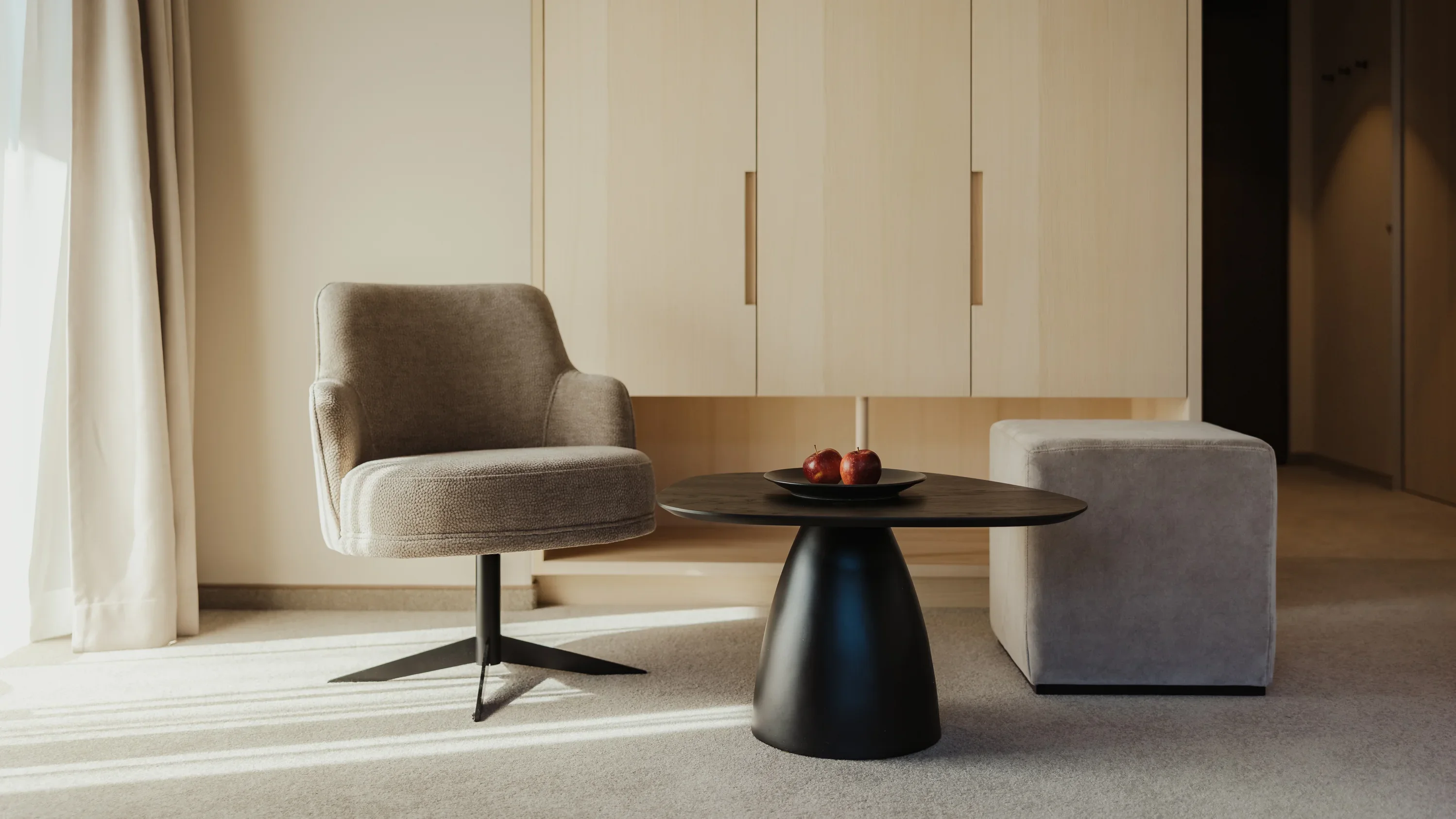 Ein modern eingerichtetes Wohnzimmer mit einem beigen Sessel, einem runden, schwarzen Tisch und einem hellgrauen Hocker. Auf dem Tisch liegen rote Äpfel. Im Hintergrund befindet sich ein heller Holzschrank. Licht fällt durch ein Fenster mit Vorhängen.