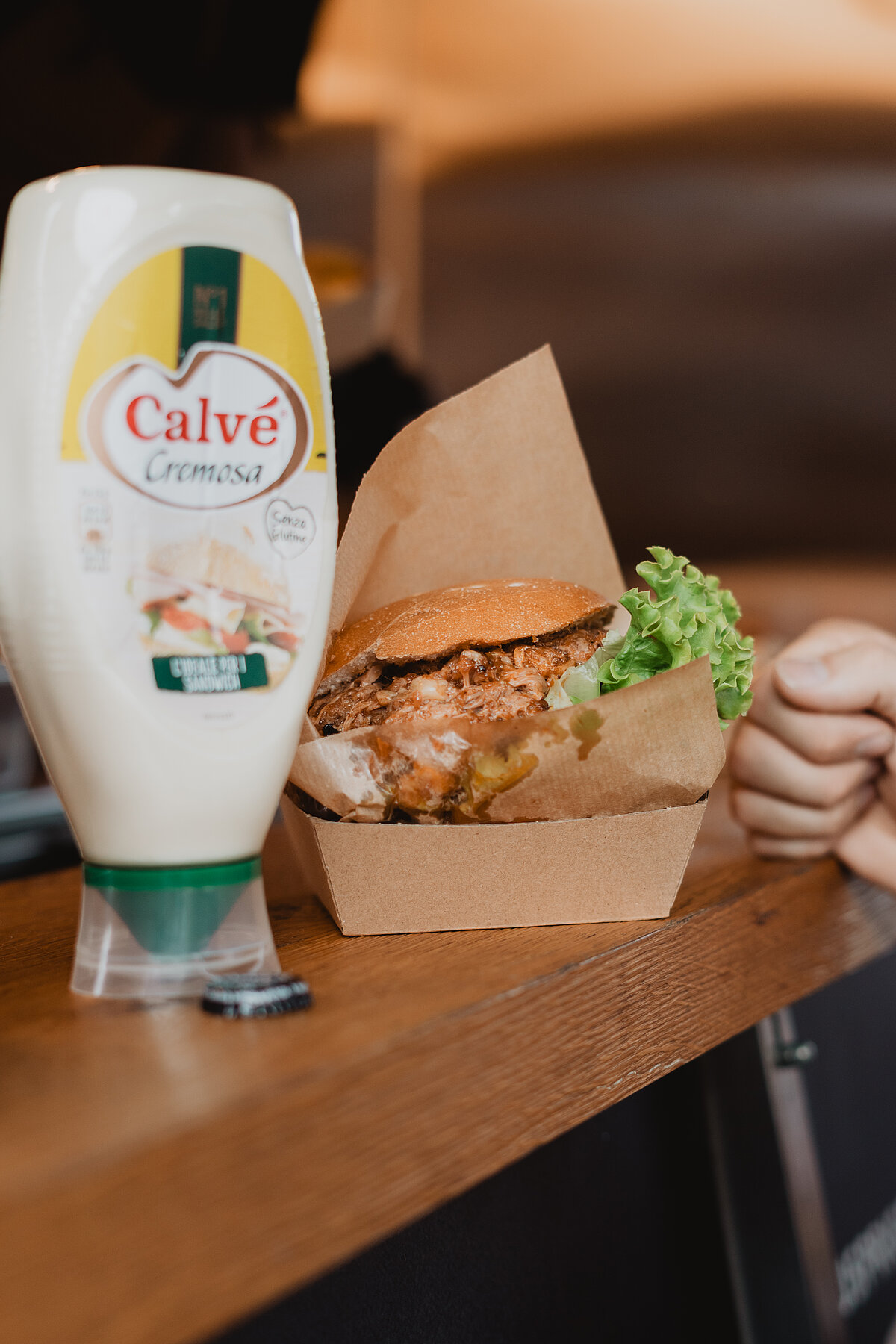 Eine Nahaufnahme eines Burgers in einer Pappschachtel auf einer Theke. Daneben steht eine Flasche Calvé Cremosa Mayonnaise. Im Hintergrund ist eine Person mit der Hand zu sehen.