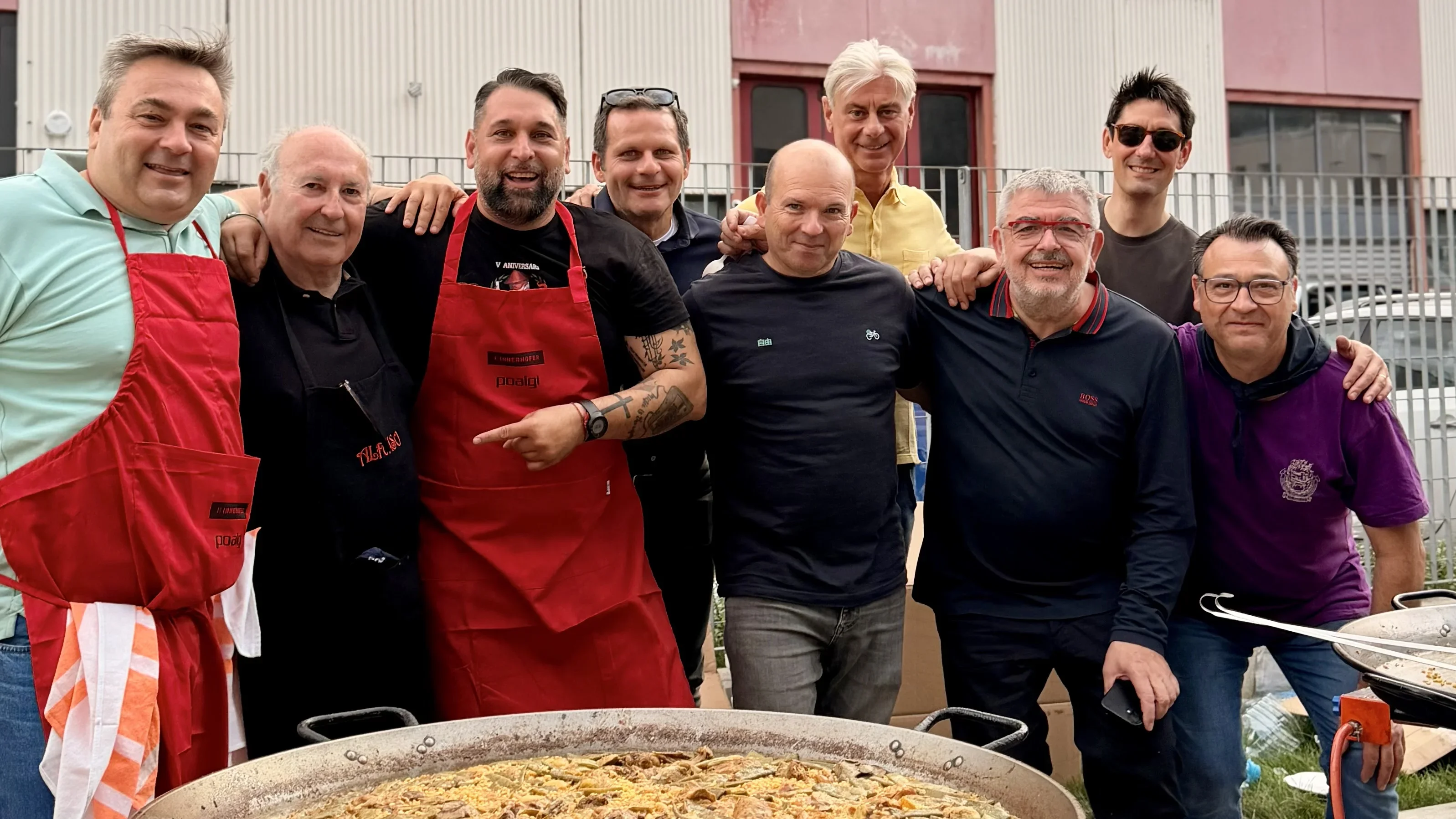 Eine Gruppe von neun Männern steht lächelnd hinter einer großen Paella-Pfanne. Einige tragen bunte Schürzen. Sie befinden sich draußen vor einem Gebäude mit rosa und weißen Wänden. Der Himmel ist bewölkt.