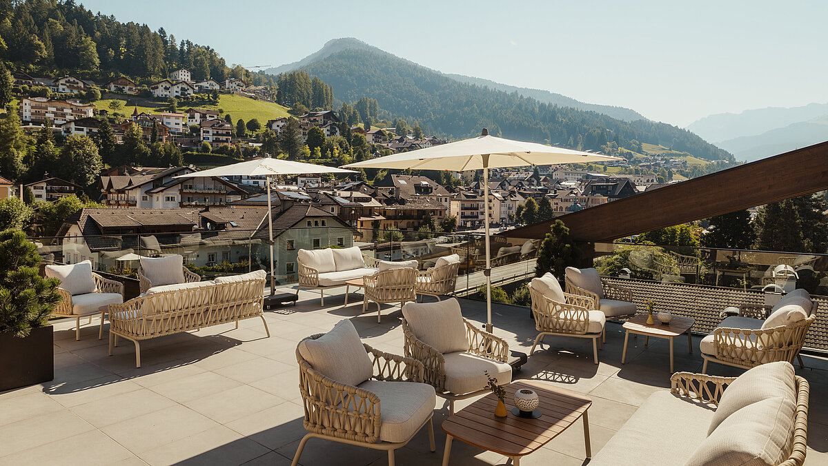 Eine Terrasse mit gemütlichen Rattansesseln und Tischen, beschattet durch Sonnenschirme. Im Hintergrund eine malerische Berglandschaft und ein Dorf mit verstreuten Häusern auf einem grünen Hügel. Der Himmel ist klar und sonnig.