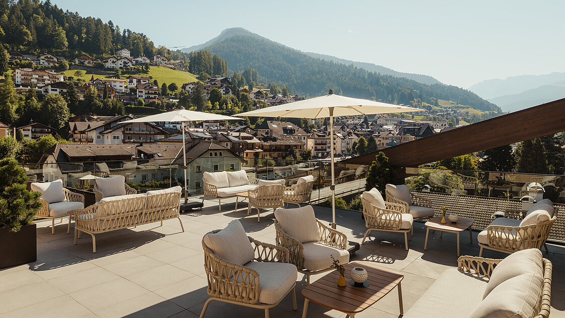 Eine Terrasse mit gemütlichen Rattansesseln und Tischen, beschattet durch Sonnenschirme. Im Hintergrund eine malerische Berglandschaft und ein Dorf mit verstreuten Häusern auf einem grünen Hügel. Der Himmel ist klar und sonnig.