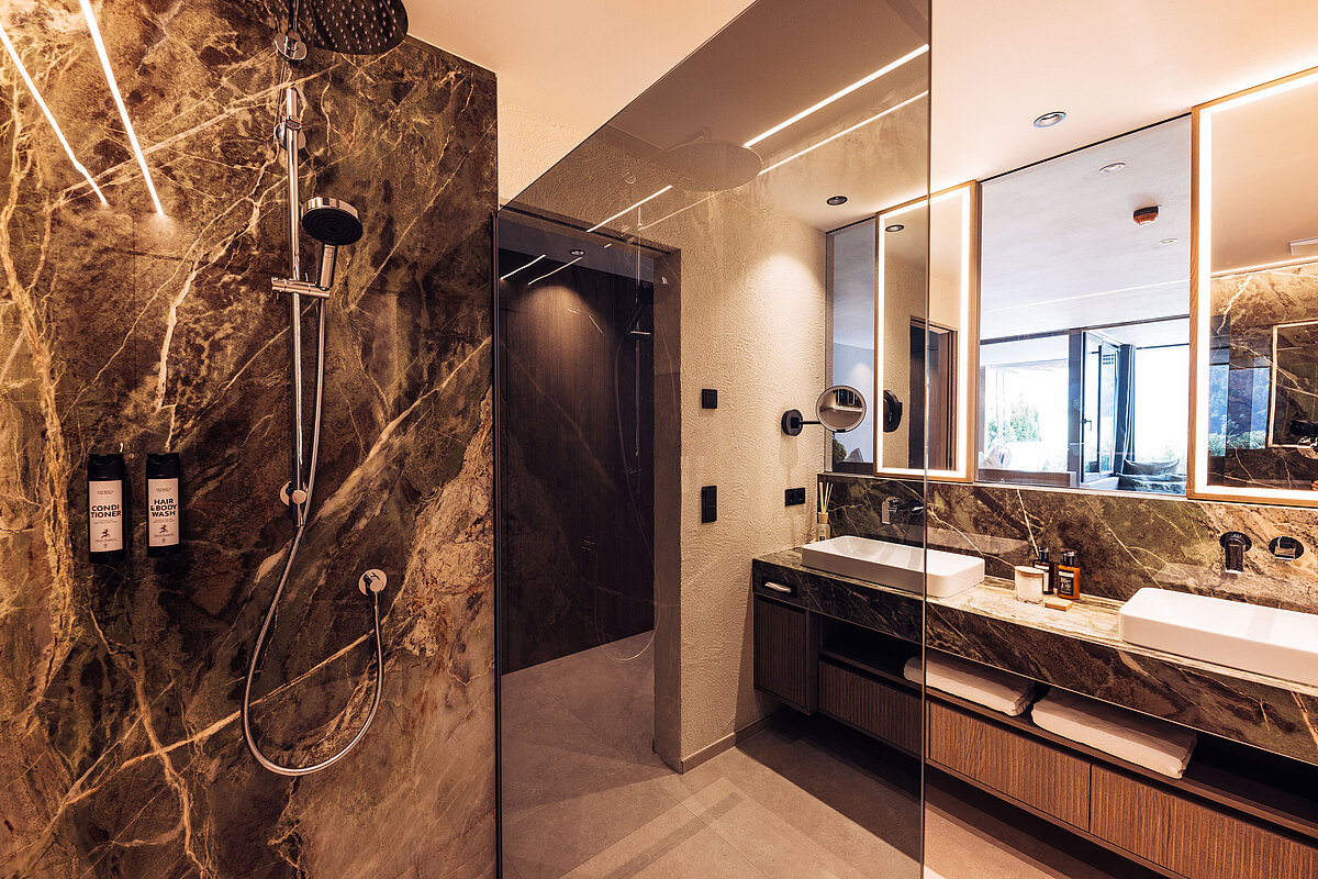 Luxuriöses Badezimmer mit eleganter Marmordusche, modernem Doppelwaschtisch mit großen Spiegeln und dezenter Beleuchtung. Der Raum strahlt ein hochwertiges Ambiente aus und enthält Handtücher, Pflegeprodukte und durch ein Fenster ist ein Außenbereich sichtbar.