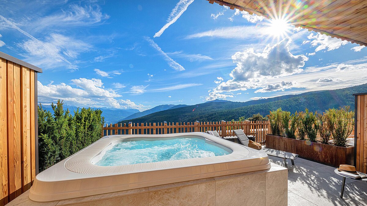 Ein Whirlpool auf einer Terrasse mit Blick auf eine malerische Berglandschaft unter einem klaren, sonnigen Himmel. Holzwände und Pflanzen im Hintergrund schaffen eine einladende und entspannende Atmosphäre.