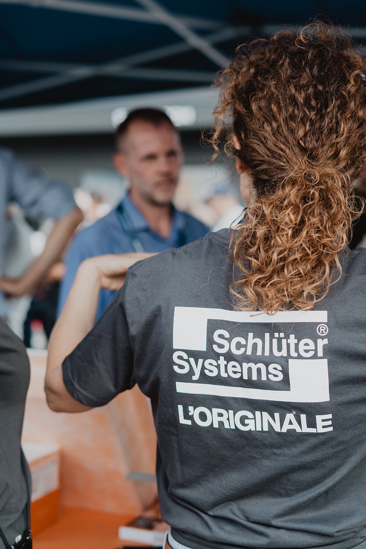 Person mit Locken trägt ein T-Shirt mit dem Aufdruck "Schlüter Systems L'ORIGINALE". Im Hintergrund unterhält sich eine Gruppe von Menschen bei einer Art von Veranstaltung.