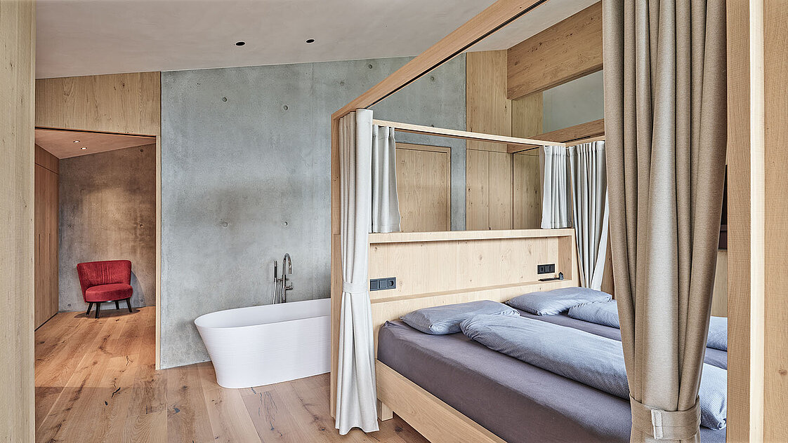Modernes Schlafzimmer mit Himmelbett aus Holz und grauer Bettwäsche. Im Raum befindet sich eine freistehende weiße Badewanne. Der Boden ist aus Holz, die Wände sind grau und minimalistisch gestaltet. Im Durchgang steht ein roter Sessel.