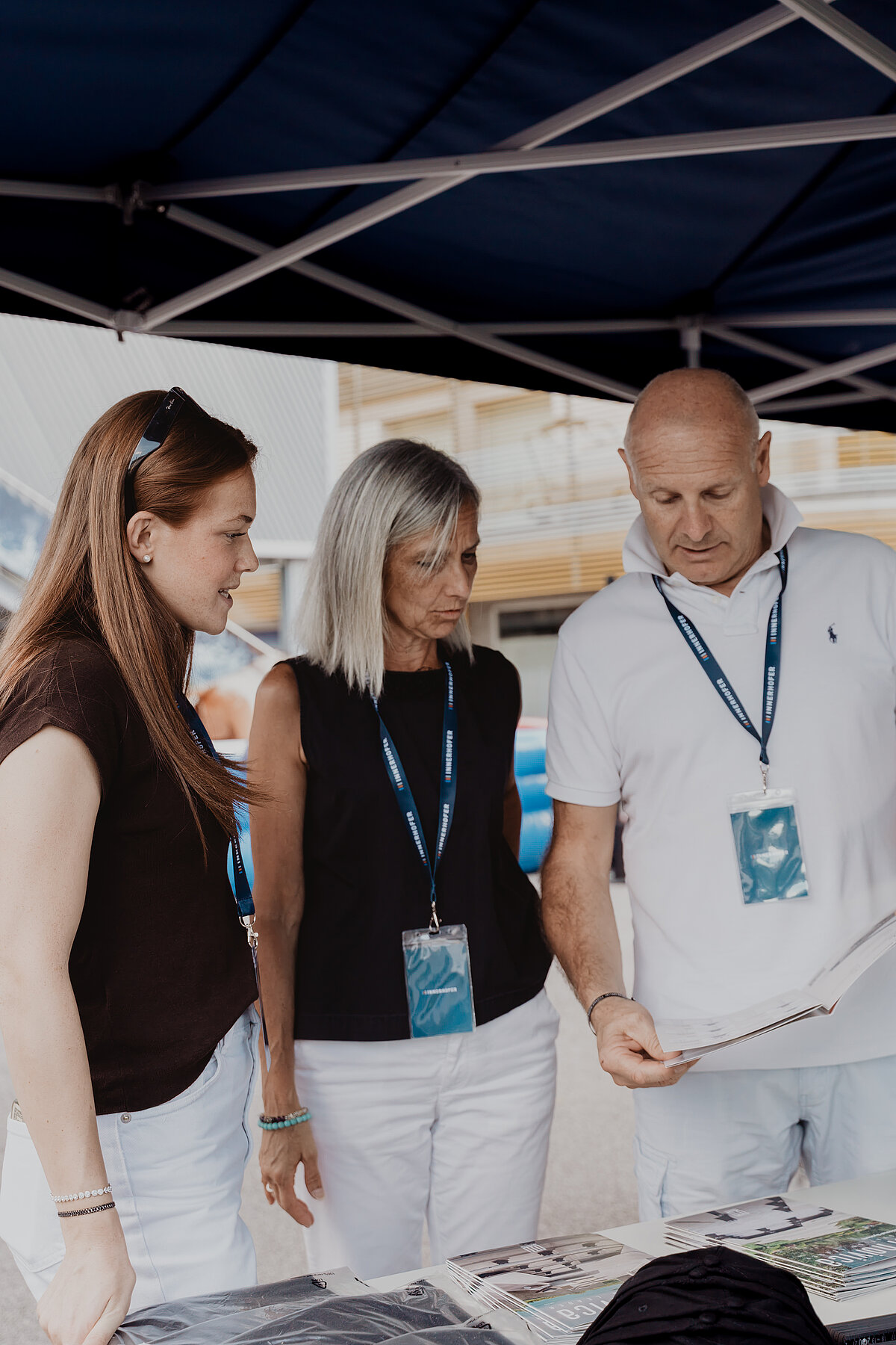 Drei Personen stehen unter einem blauen Pavillon und betrachten gemeinsam ein Blatt Papier. Alle tragen dunkle Oberteile und helle Hosen, und sie haben ID-Karten um den Hals. Im Hintergrund ist ein unscharfes Gebäude zu sehen.