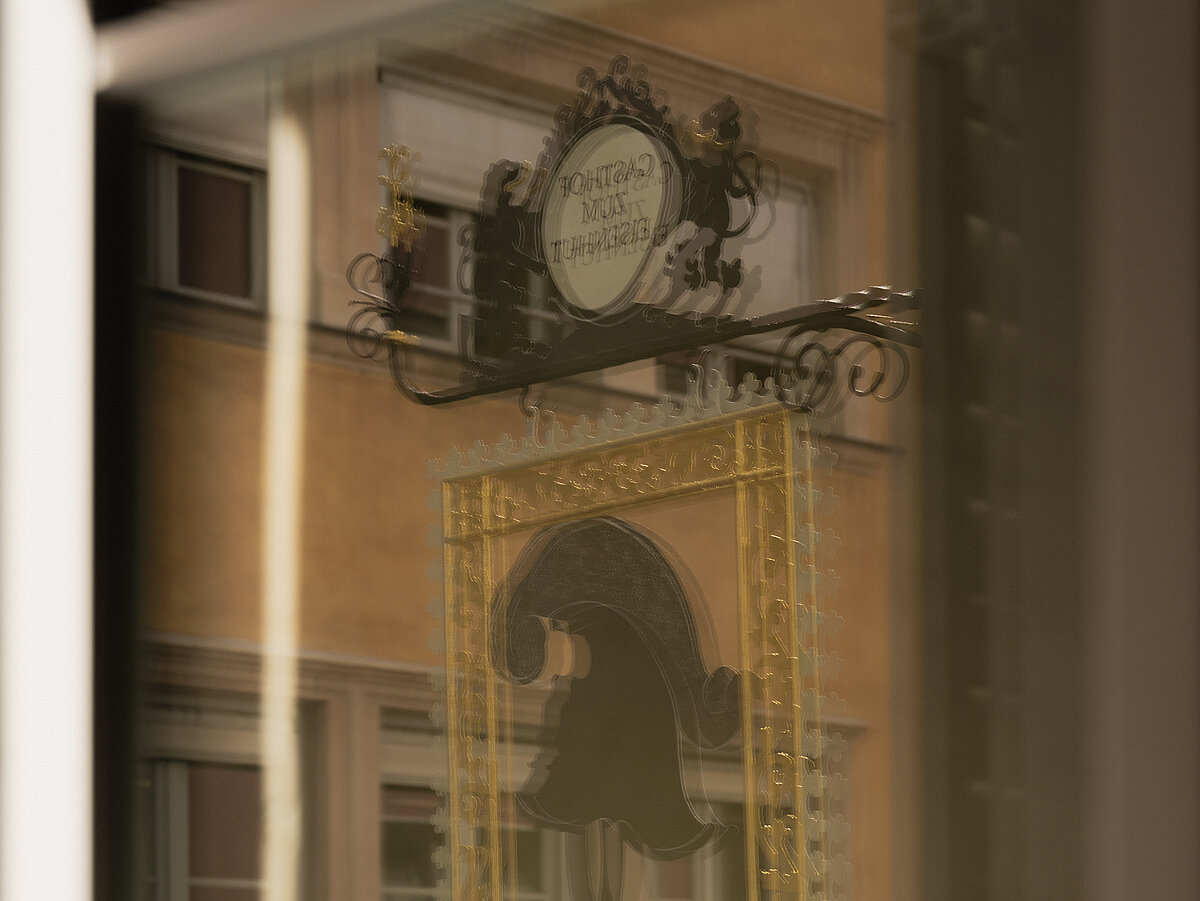 Spiegelung eines traditionellen hängenden Wirtshaus- oder Ladenschilds mit kunstvoll gestalteten Elementen in einem Fenster, durch das eine beige Gebäudeansicht sichtbar ist. Das Schild zeigt dekorative Verzierungen und liegt vor einem unscharfen städtischen Hintergrund.