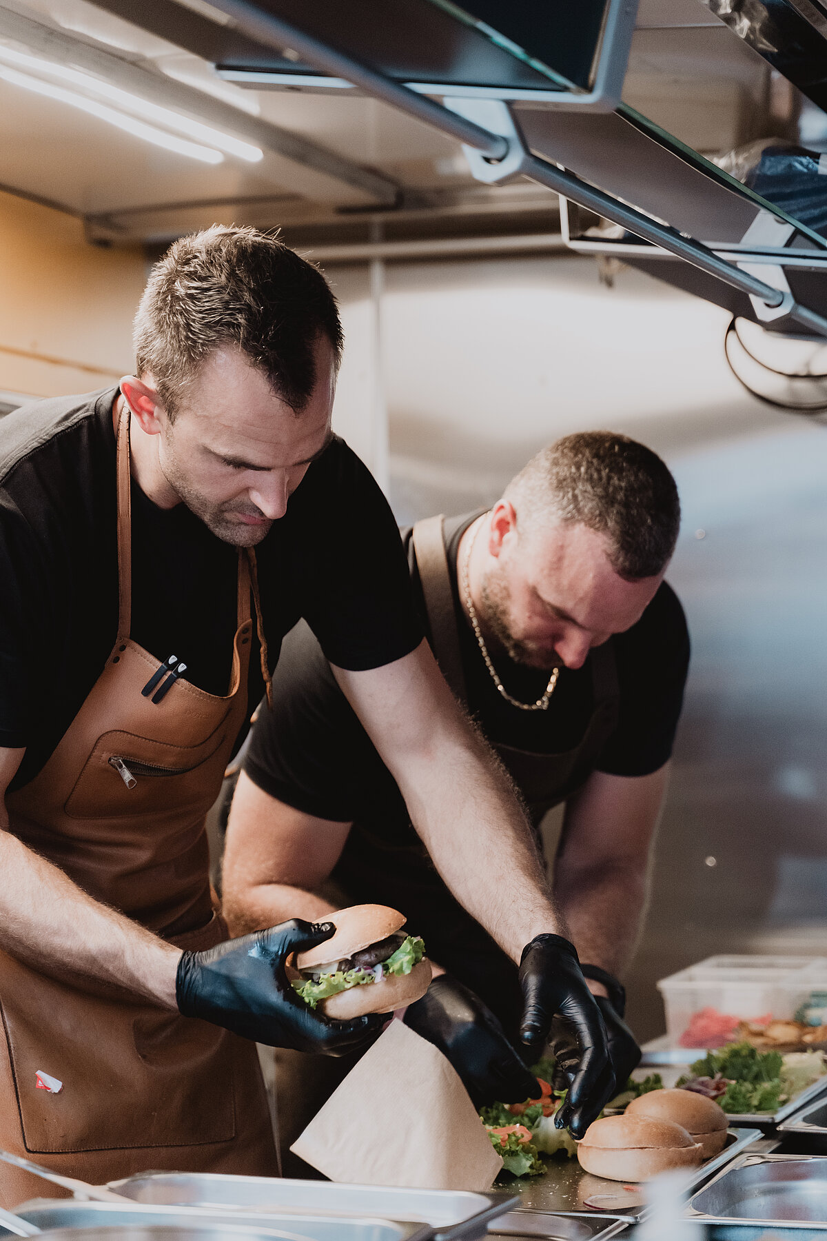 Zwei Männer in braunen Schürzen und schwarzen Handschuhen bereiten in einer Küche Hamburger zu. Einer hält einen Burger, während der andere Gemüse arrangiert. Die Umgebung ist eine professionelle Küche mit Arbeitsfläche und Zutaten.
