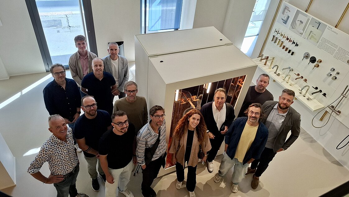 Eine Gruppe von fünfzehn Personen steht in einem modernen Ausstellungsraum rund um ein großes, quadratisches Möbelstück. An den Wänden hängen verschiedene Bad- und Küchenarmaturen. Die Personen blicken lächelnd zur Kamera.