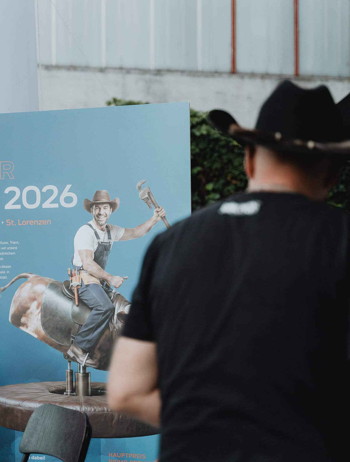 Ein Mann mit Cowboyhut steht vor einem Plakat, das einen Mann zeigt, der in Cowboykleidung auf einem mechanischen Bullen reitet und einen großen Schraubenschlüssel hält. Auf dem Plakat steht „2026“.