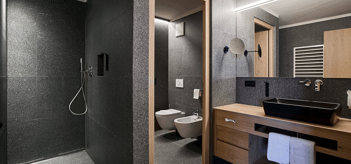 Modernes Badezimmer mit dunkler Steinwand, großem Spiegel, Holzschränken und Badewanne. Im Hintergrund ist ein Flur mit Tageslicht zu sehen.