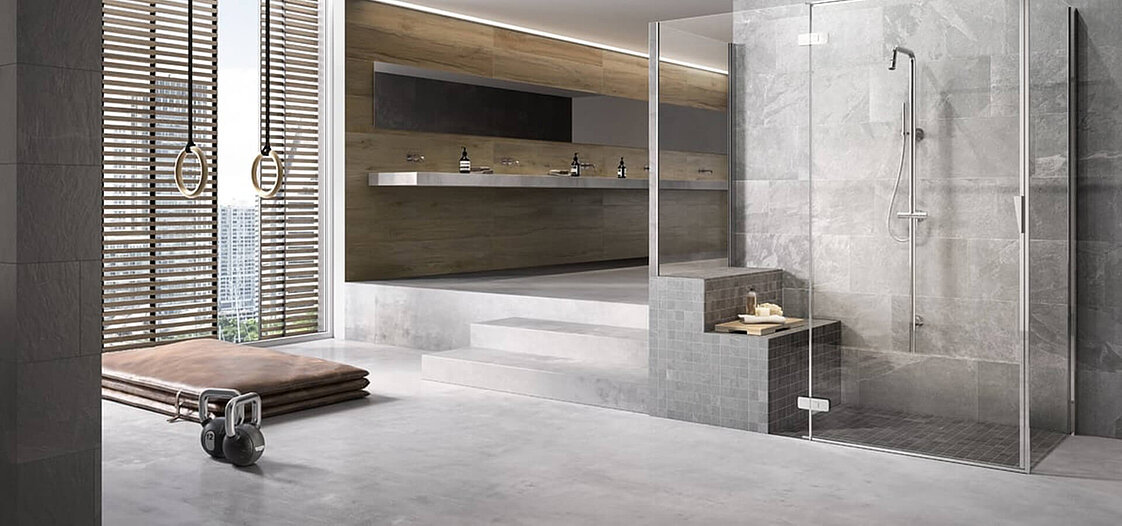 Modernes Badezimmer mit großer, verglaster Dusche, grauen Fliesen und minimalistischer Einrichtung. Im Hintergrund befinden sich Doppelwaschbecken und ein breites Spiegelpaneel. Auf der linken Seite ist eine gepolsterte Liege mit Gymnastikringen an Holzwänden sichtbar.