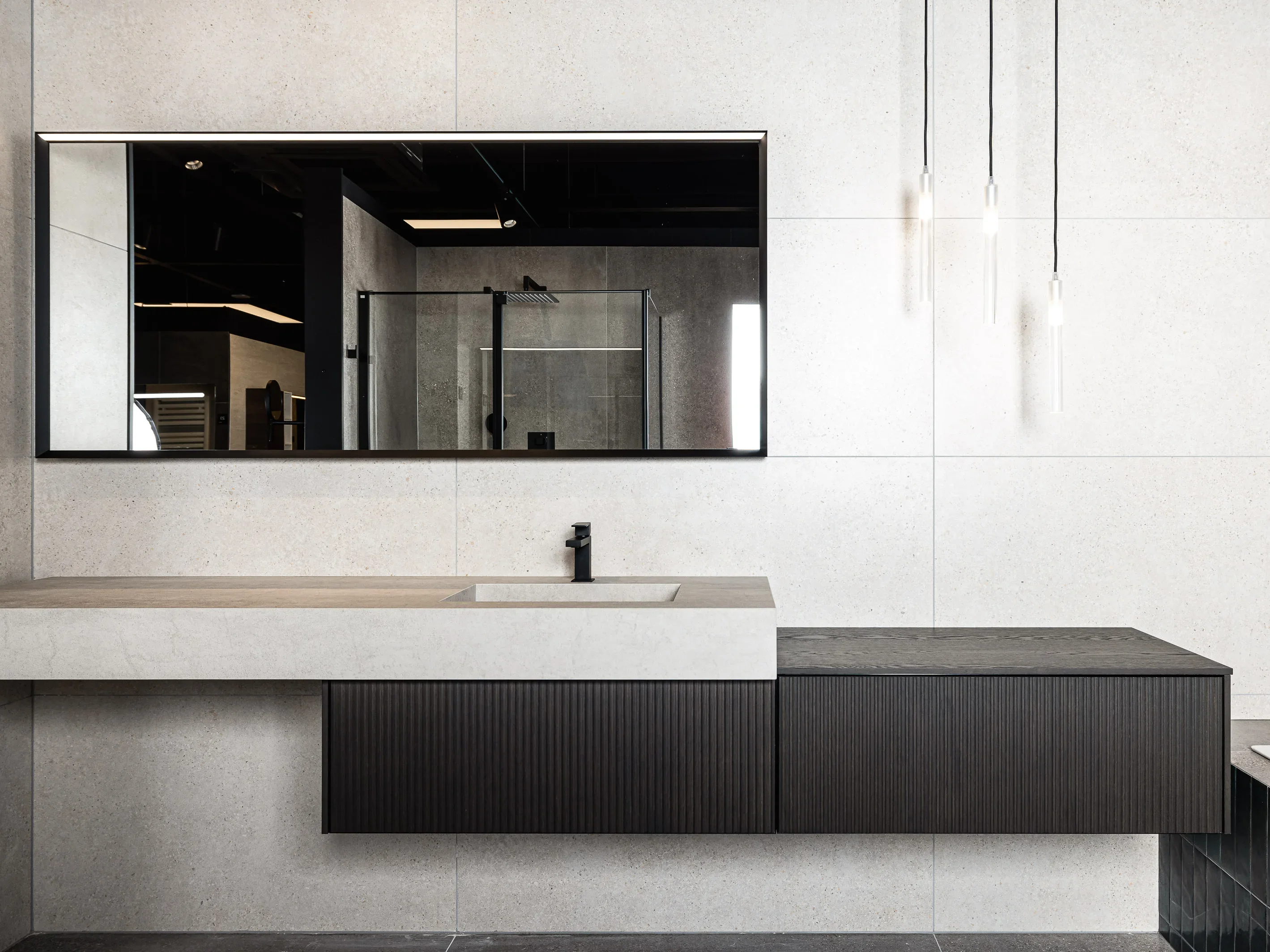 Modernes Badezimmer mit großen, rechteckigen Spiegel über einem minimalistischen Waschbecken. Dunkle, schlichte Hängeschränke und ausgefallene Hängeleuchten ergänzen das Design. Elegante, helle Wandfliesen runden die zeitgenössische Ästhetik ab.