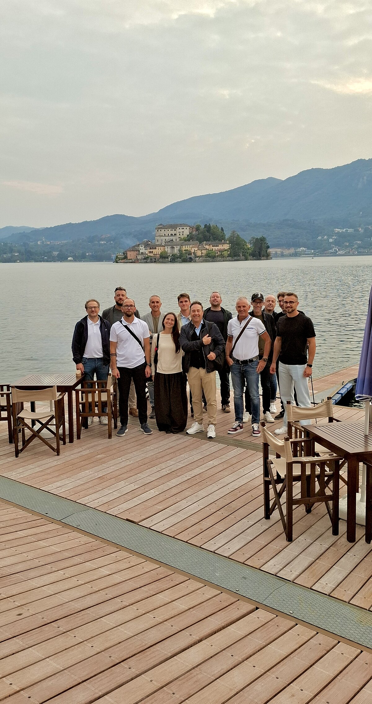 Eine Gruppe von Menschen steht auf einem Holzsteg am See, im Hintergrund eine Insel mit einem Gebäude. Der Himmel ist bewölkt, und man sieht umliegende Berge. Tische und Stühle sind auf dem Steg verteilt.