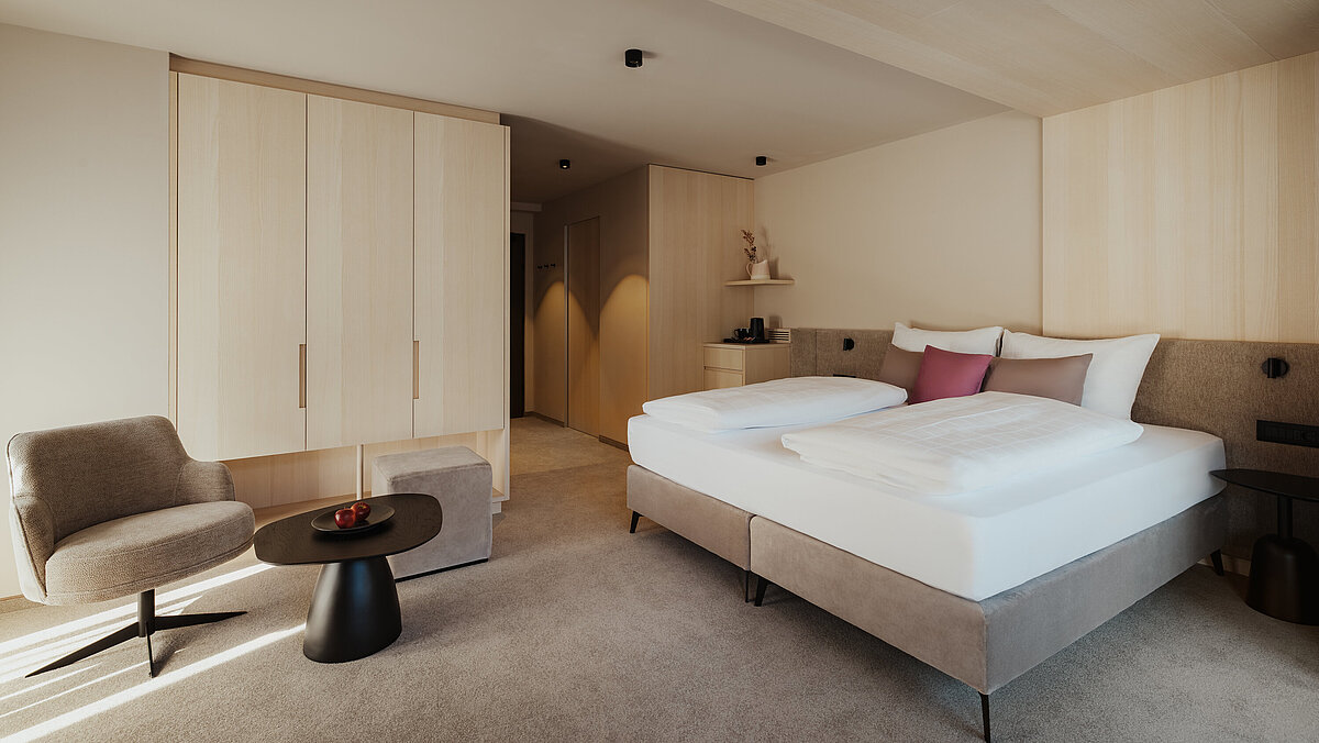 Ein modernes Hotelzimmer mit einem großen Doppelbett, flauschigen Kissen und einer beigen Tagesdecke. Neben dem Bett steht ein bequemer Sessel. Ein kleiner, runder Tisch mit einer Dekoration darauf ergänzt das gemütliche Ambiente. Helle Holzmöbel und Teppichboden dominieren den Raum.