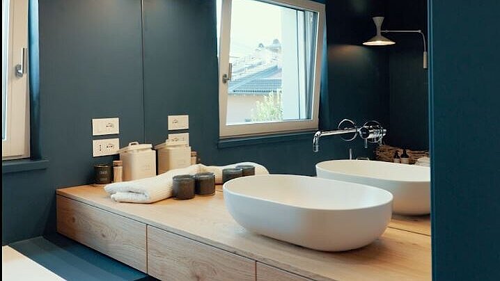 Modernes Badezimmer mit blau gestrichenen Wänden, Holzdekor und beleuchtetem Spiegel. Ein weißes Waschbecken steht auf einer Holztischplatte, daneben sind Pflegeprodukte. Ein Fenster lässt natürliches Licht ein.