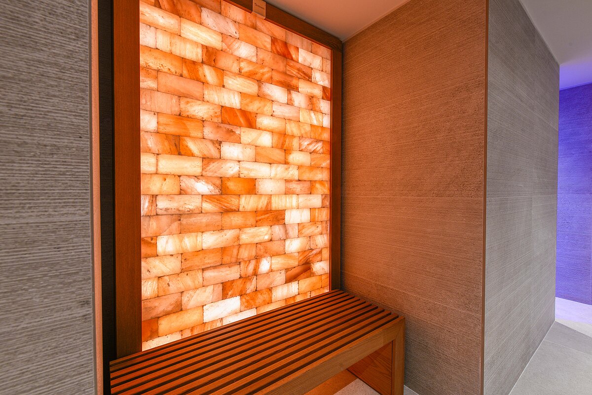 Ein beleuchtetes Salzziegelmauerwerk hinter einer hölzernen Sitzbank in einem modernen Spa-ähnlichen Raum. Die Mauer besteht aus orangefarbenen Salzziegeln, die warmes Licht ausstrahlen.