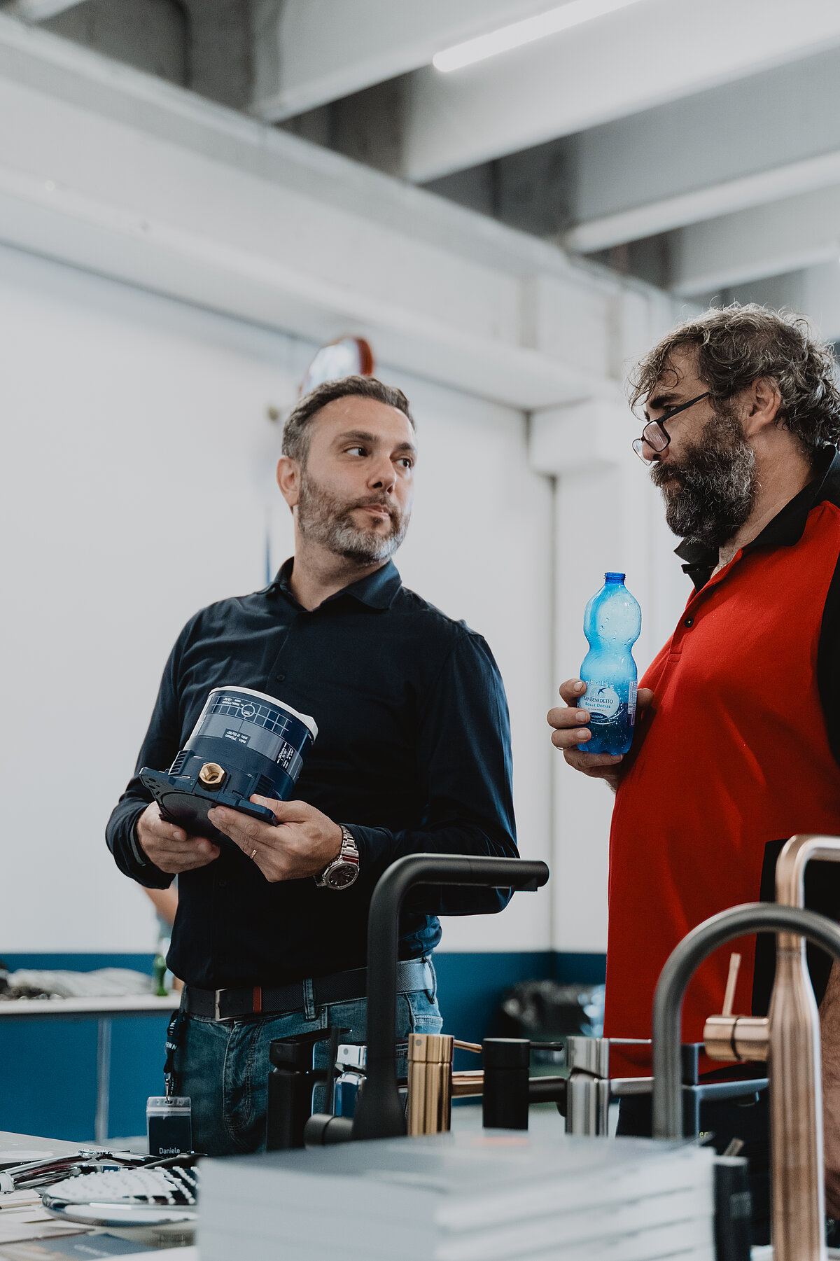 Zwei Männer in einem industriellen Raum, diskutieren. Einer hält eine blaue Verpackung, der andere eine blaue Wasserflasche. Vor ihnen liegen Metallgegenstände auf einem Tisch.
