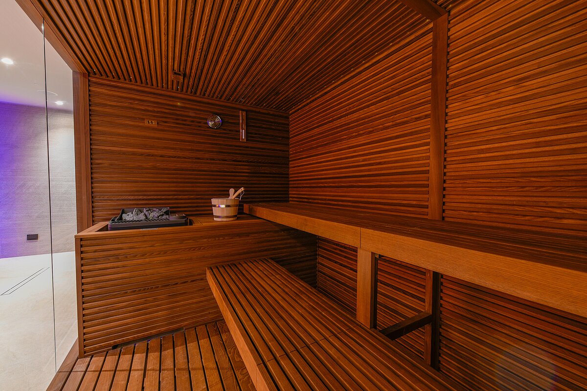 Holzverkleidete Sauna mit Bank und Saunaofen. Ein hölzerner Eimer mit Kelle steht neben dem Ofen. Links ist eine Glastür, die zum beleuchteten Raum führt.