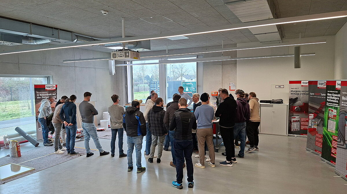 Eine Gruppe von Menschen steht in einem modernen Raum und hört einem Redner zu, der technische Ausrüstung präsentiert. Informationsbanner sind im Hintergrund sichtbar.