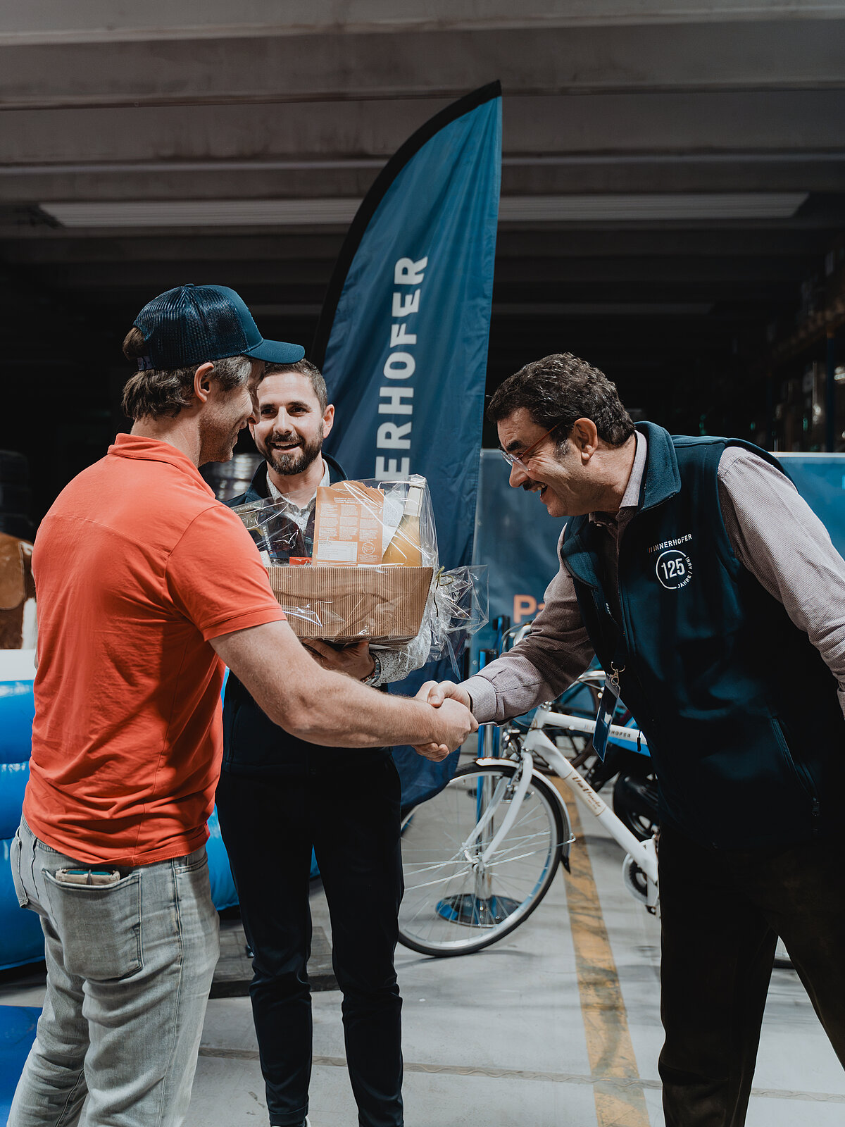 Drei Männer stehen in einer Garage. Einer trägt ein rotes Hemd und schüttelt die Hand eines Mannes in einer Weste. Ein weiterer Mann hält einen Geschenkkorb und lächelt. Im Hintergrund ist ein blaues Banner und ein Fahrrad zu sehen.
