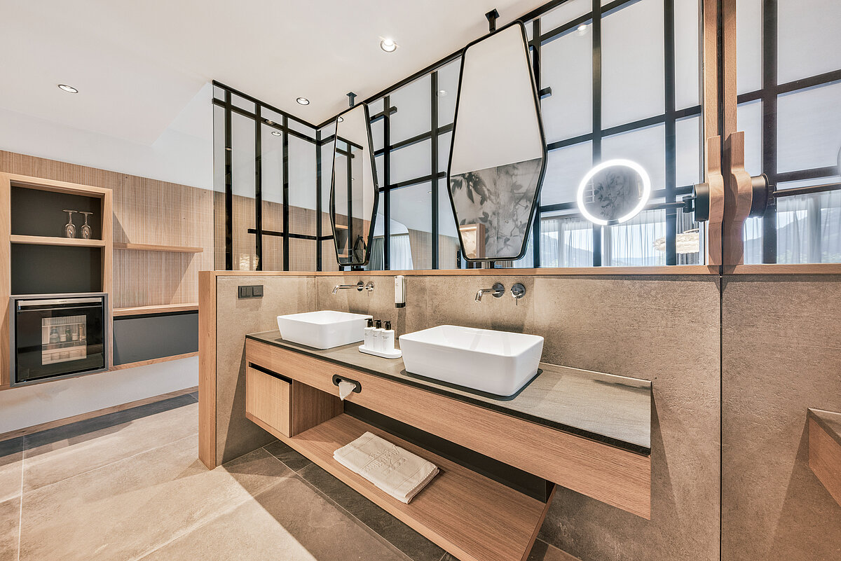 Modernes Badezimmer mit zwei eckigen Waschbecken, großen Spiegeln und einem Holzregal darunter. Die Rückwand besteht aus dunklem Glas und einem quadratischen Wandregal. Die Beleuchtung ist dezent und geliefert durch eingebaute Deckenleuchten.