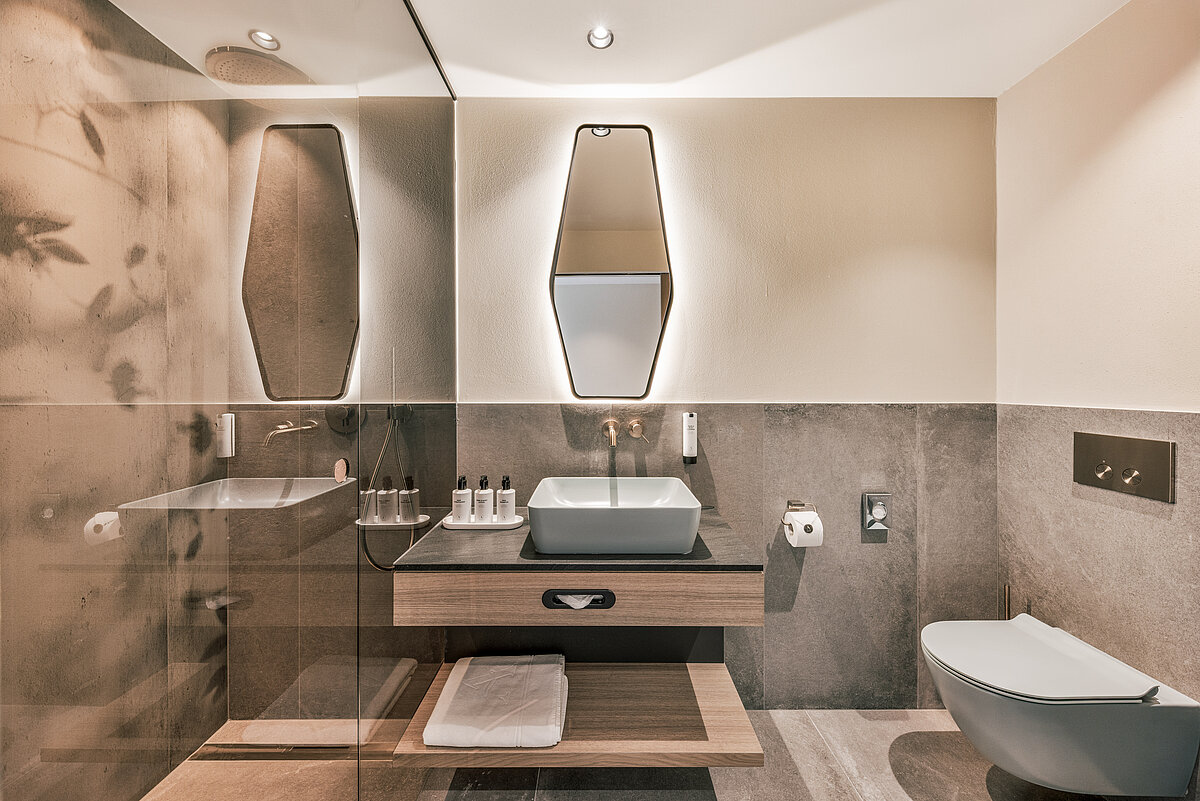 Modernes Badezimmer mit großem Spiegel über einem rechteckigen Waschbecken auf einem hölzernen Waschtisch. Neben dem Waschbecken sind Hygieneartikel angeordnet. Rechts befindet sich eine schwebende Toilette. Das Design kombiniert neutrale Töne und elegante Beleuchtung.