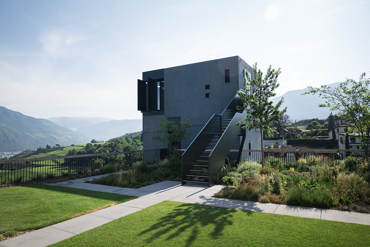 Ein modernes, kubisches Gebäude mit einer äußeren Treppe steht in einer grünen, landschaftlich gestalteten Umgebung, umgeben von Rasenflächen und Bäumen. Im Hintergrund sind Hügel und Berge unter einem klaren Himmel sichtbar.