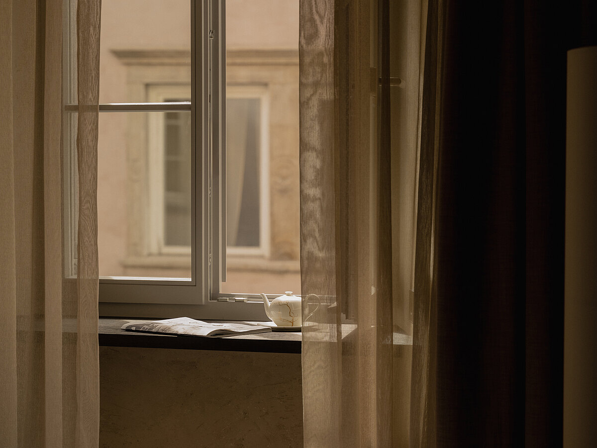 Ein halb geöffneter Vorhang aus dünnem Stoff lässt Tageslicht durch ein Fenster in einem ruhigen Raum. Auf der Fensterbank steht eine Tasse mit Untertasse. Das Fenster bietet einen Blick auf eine gegenüberliegende Hauswand. Im Vordergrund ist eine Ecke eines Bettes sichtbar.