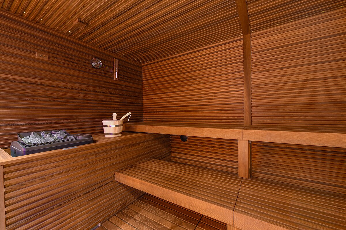 Innenansicht einer holzverkleideten Sauna mit Holzbank und Saunaofen. Ein hölzerner Aufgusseimer mit Schöpflöffel steht auf der Bank. Die Sauna ist leer und zeigt eine gemütliche, warme Atmosphäre.
