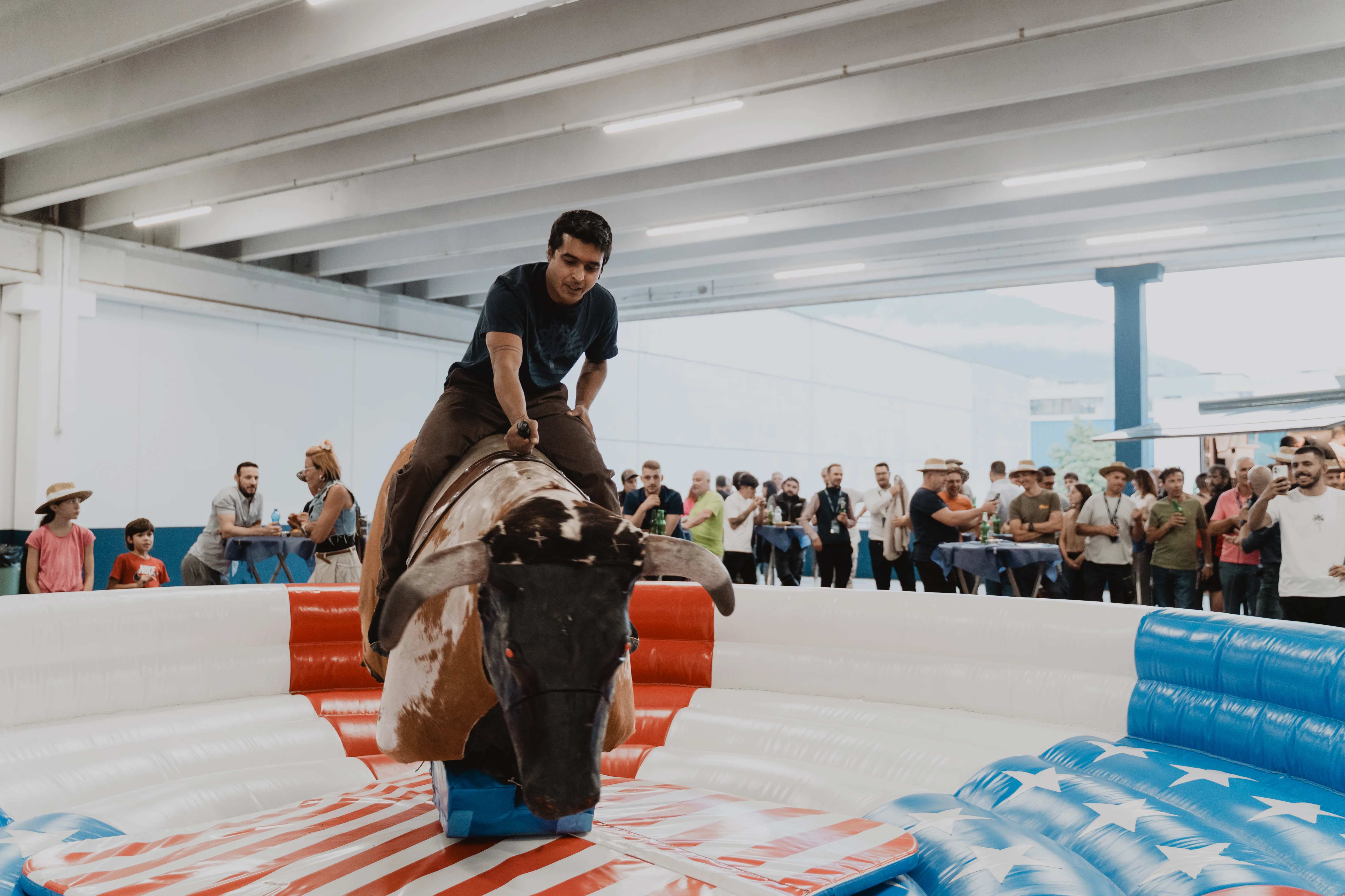 Ein Mann reitet auf einem mechanischen Bullen in einer Indoor-Anlage. Der Bullenring ist mit amerikanischen Flaggenmotiven verziert. Zuschauer stehen im Hintergrund und beobachten das Geschehen.