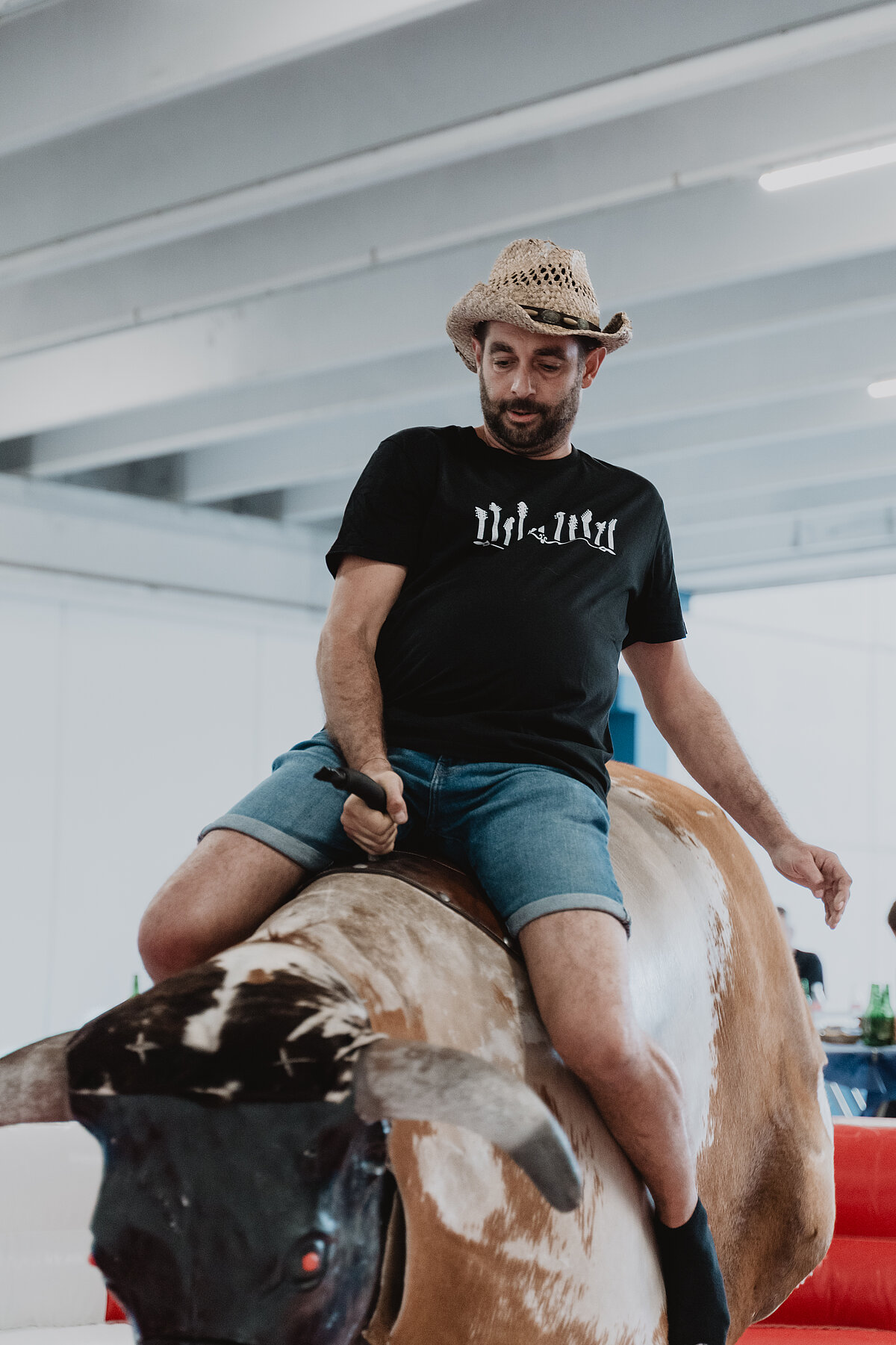 Ein Mann trägt einen Strohhut und ein schwarzes T-Shirt, während er auf einem mechanischen Bullen in einer Halle reitet. Er hält sich mit einer Hand am Bullen fest.