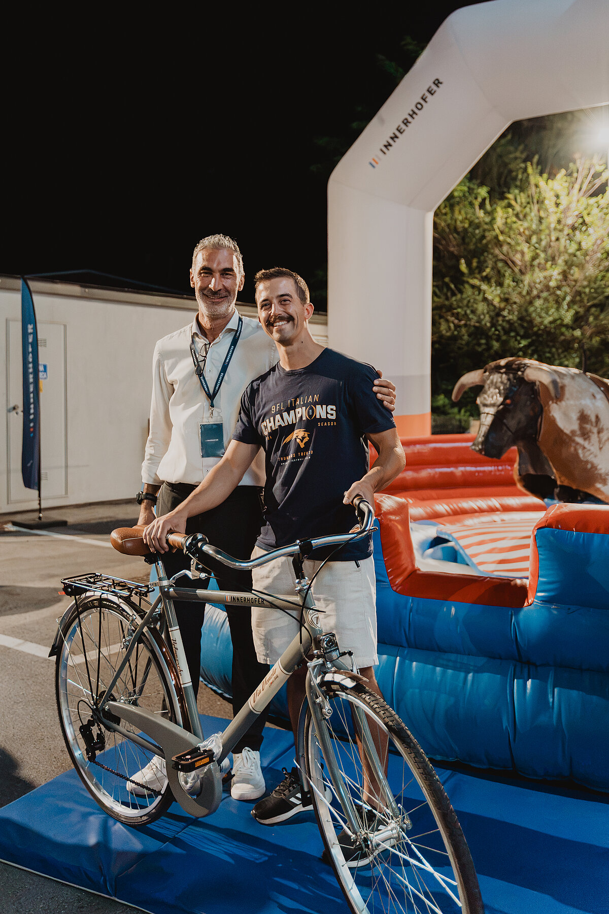 Zwei Männer lächeln und posieren für ein Foto. Der vordere Mann hält ein Fahrrad. Im Hintergrund ist ein mechanischer Bulle und eine aufblasbare Attraktion zu sehen. Es ist Nacht und die Szene ist gut beleuchtet.