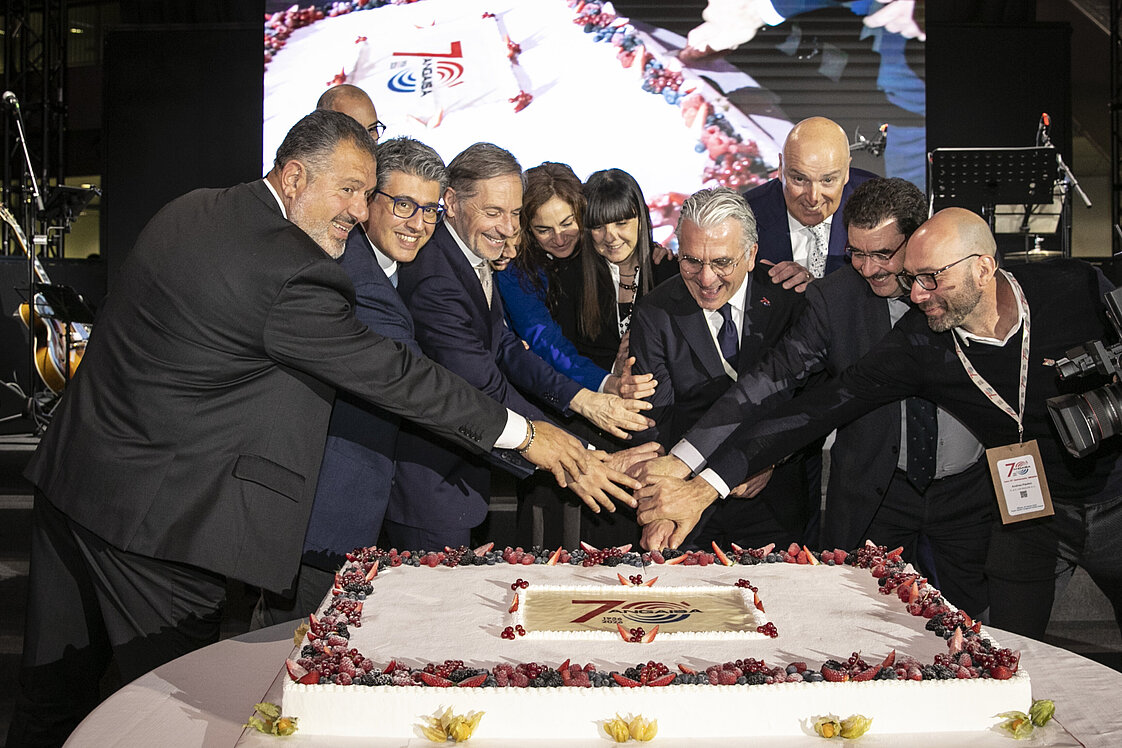 Eine Gruppe von Menschen steht um eine große rechteckige Torte mit roten Verzierungen, die das Logo "75" zeigt. Sie lächeln und halten gemeinsam Messer zum Anschneiden der Torte. Im Hintergrund ist ein Bildschirm mit einer größeren Darstellung der Torte zu sehen.