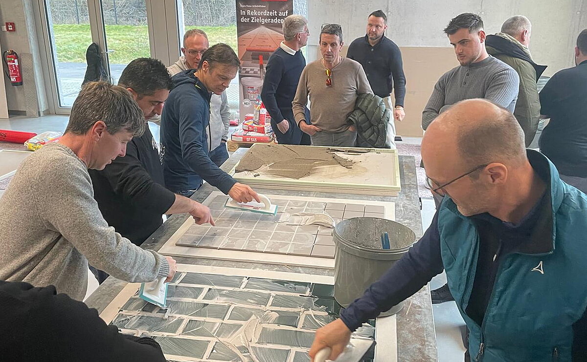 Eine Gruppe von Menschen arbeitet gemeinsam an einem Tisch. Sie tragen Fliesen oder ähnliche Materialien auf, scheinbar in einem handwerklichen oder schulischen Kontext. Alle konzentrieren sich auf die Aufgabe und benutzen Werkzeuge, um das Material gleichmäßig zu verteilen.