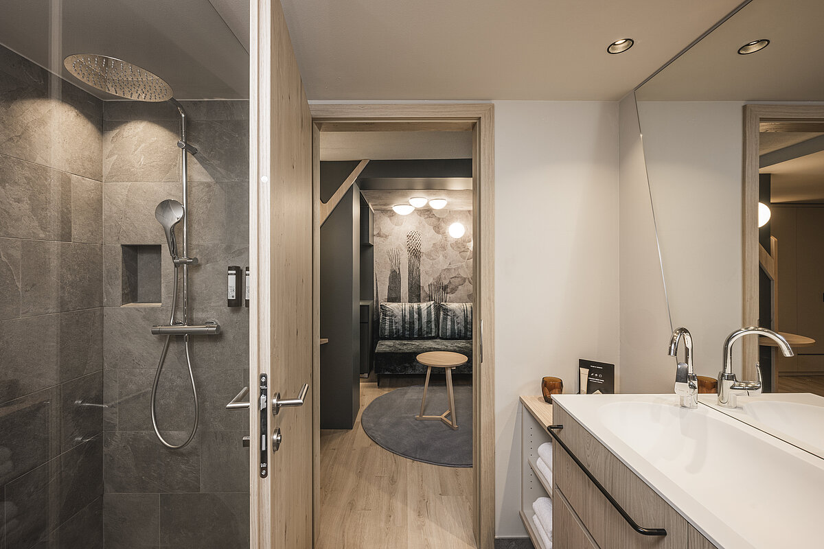 Ein modernes Badezimmer mit einer geräumigen Dusche aus dunklen Fliesen, daneben ein Waschbecken mit großem Spiegel. Im Hintergrund ist ein Wohnbereich mit einem kleinen Tisch und Sofa sichtbar, beleuchtet von mehreren Deckenlampen.