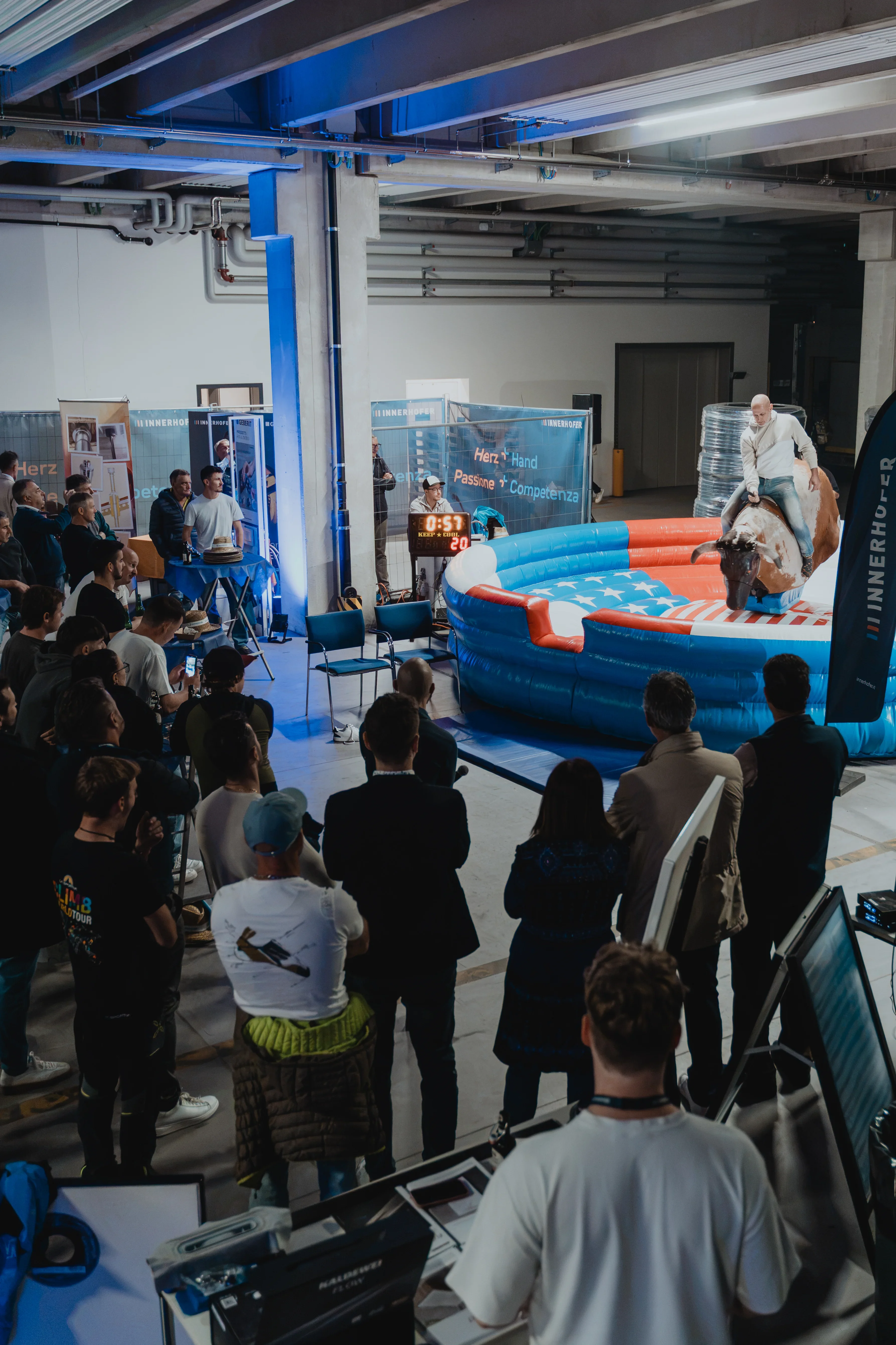 Personenmenge schaut einem Mann auf einem mechanischen Bullen in einer Indoor-Arena zu. Der Raum ist industrial gestaltet, mit blauer Akzentbeleuchtung. Technikpersonal steht in der Nähe an einem Mischpult.