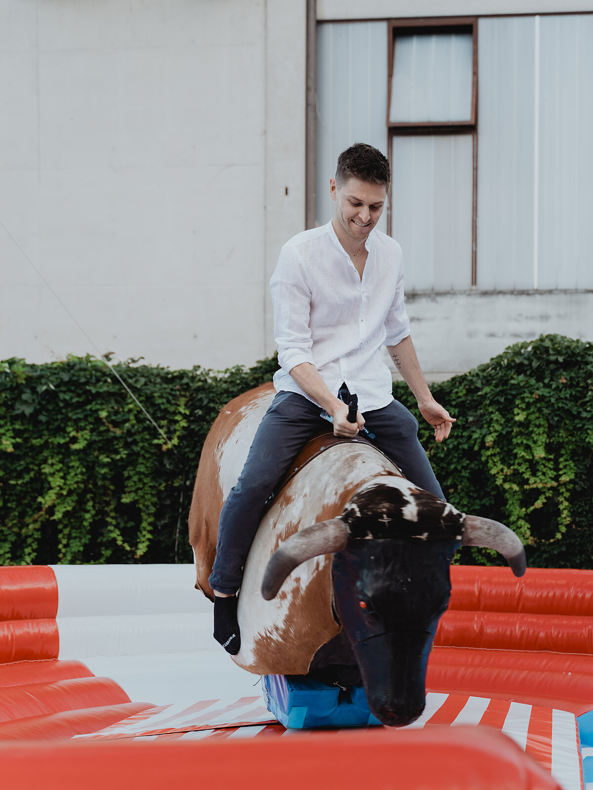 Ein Mann mit weißem Hemd reitet auf einem mechanischen Bullen in einer aufblasbaren Arena. Im Hintergrund sind grüne Pflanzen und eine Gebäudefassade sichtbar.