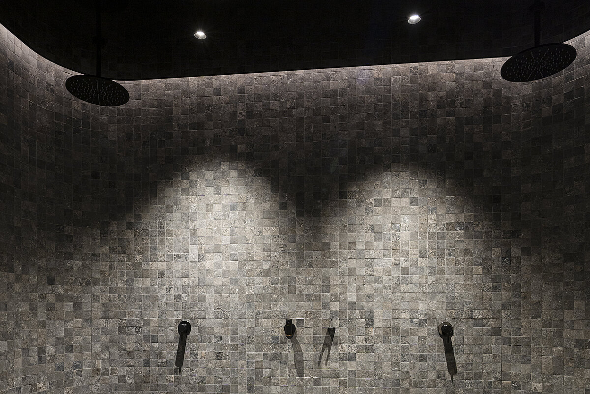 Dusche in einem modernen Badezimmer, mit grauen Fliesen an den Wänden und dem Boden. Es sind mehrere Duschköpfe und Armaturen an der Wand angebracht. Die Beleuchtung ist dezent und schafft eine entspannte Atmosphäre.