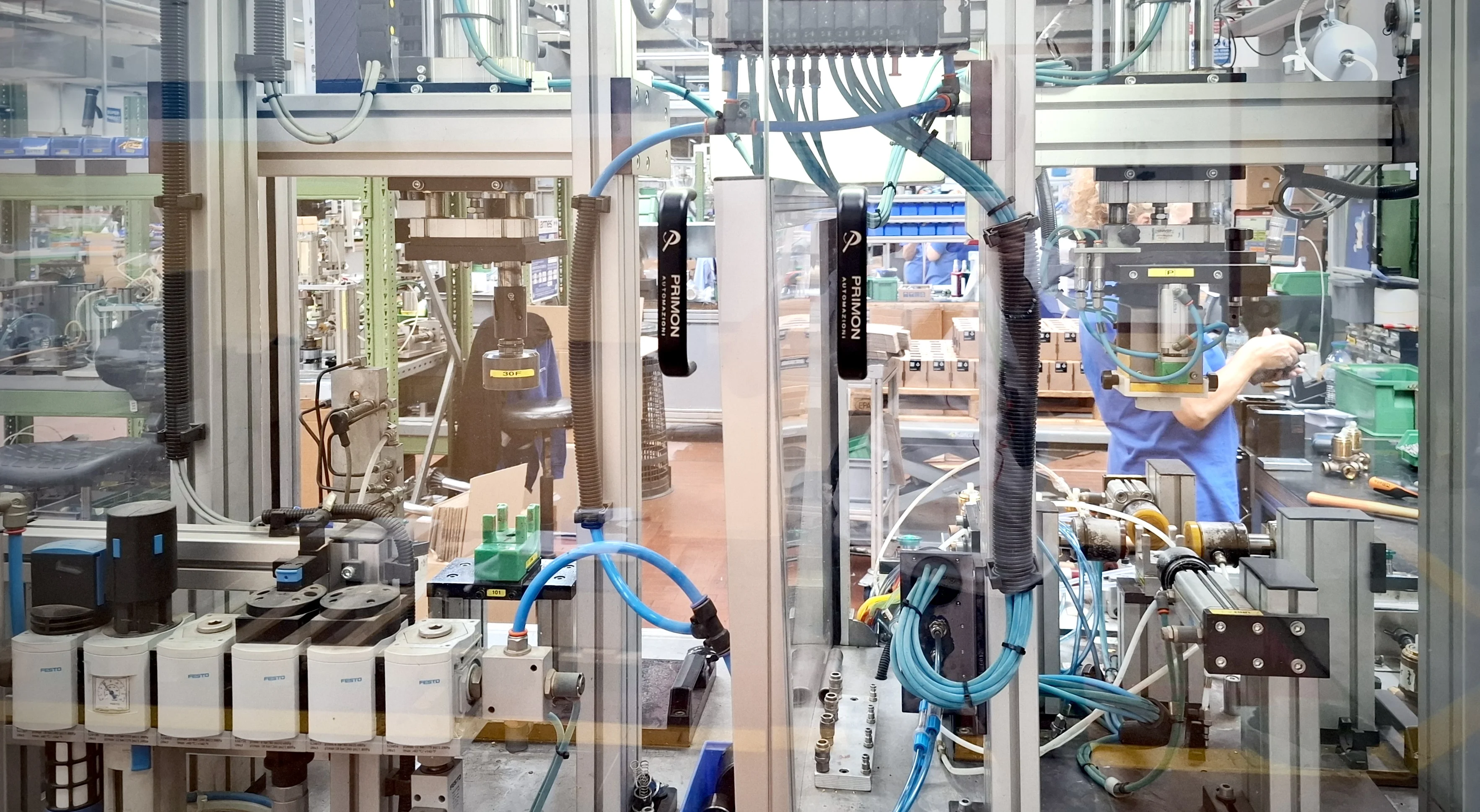 Industrielle Fabrikanlage mit moderner Automatisierungstechnik, einschließlich Roboterarmen, Kabeln und Sensoren. Im Hintergrund arbeitet eine Person an einer Maschine. Die Umgebung wirkt wie eine fortschrittliche Produktionsstätte mit komplexen technischen Geräten.