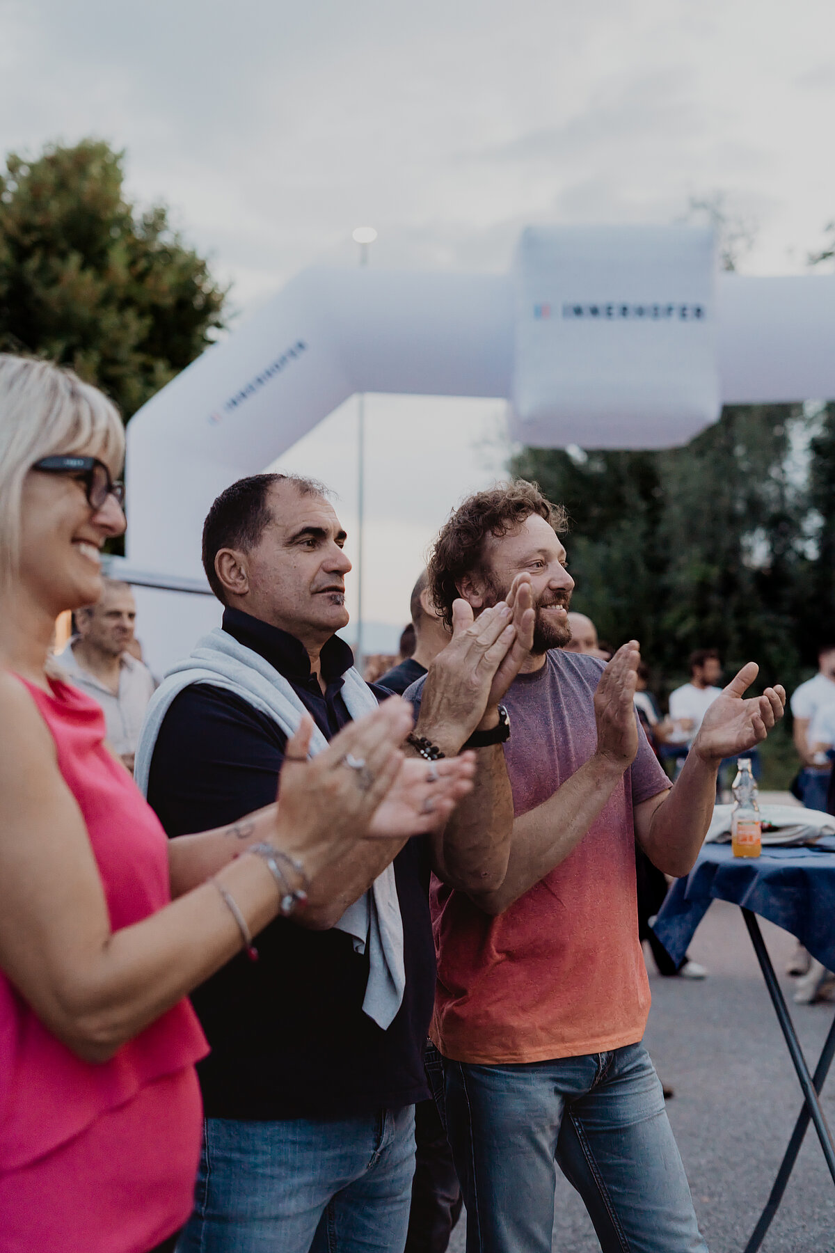 Menschen, die im Freien bei einer Veranstaltung klatschen. Eine Frau steht links und trägt eine Sonnenbrille, daneben zwei Männer, die fröhlich applaudieren. Im Hintergrund ist ein aufblasbares Tor mit der Aufschrift "Innerhofer" zu sehen.
