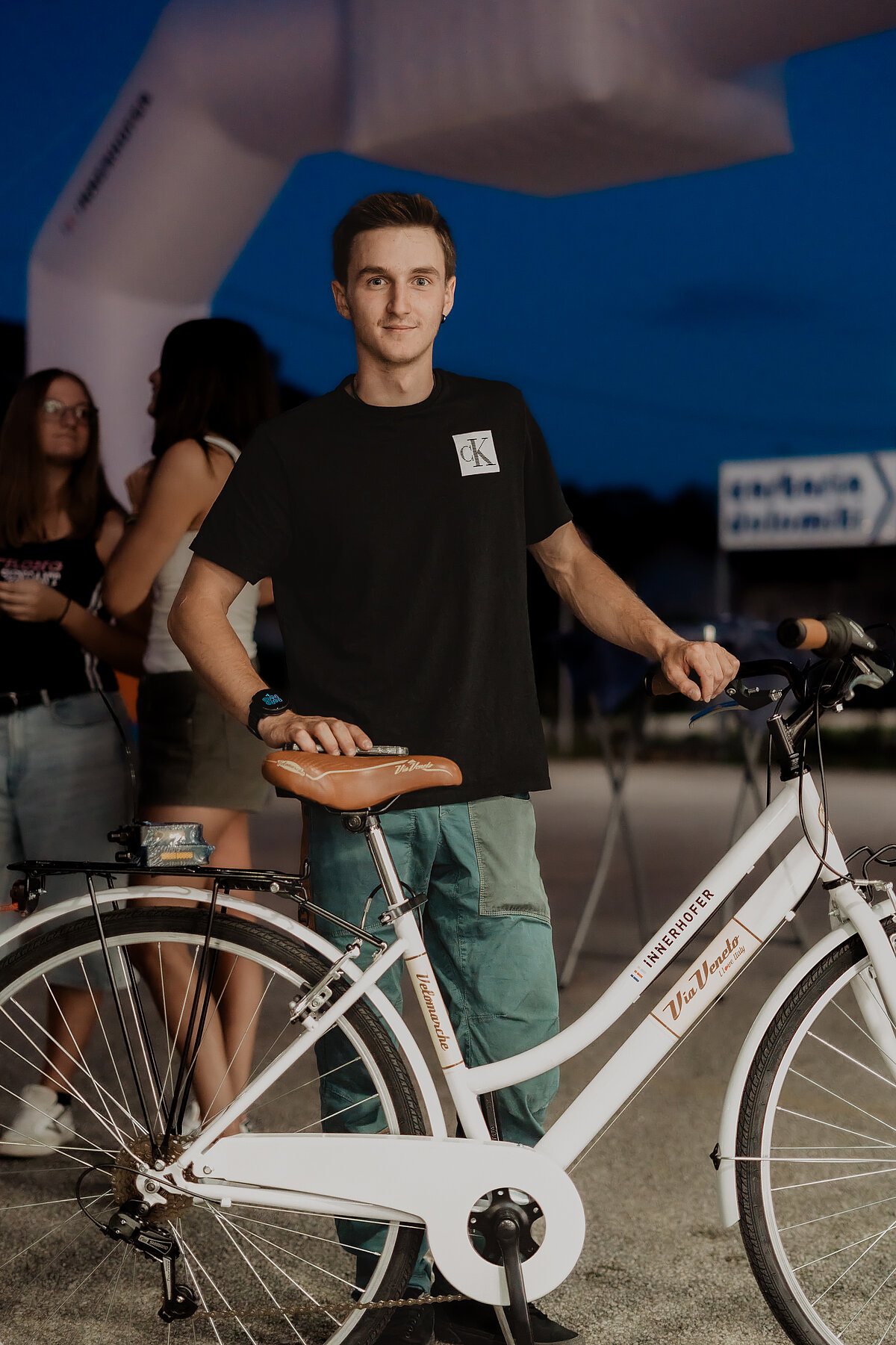 Ein Mann steht neben einem weißen Fahrrad auf einer nächtlichen Veranstaltung im Freien. Im Hintergrund sind andere Personen und ein aufblasbarer Torbogen sichtbar.