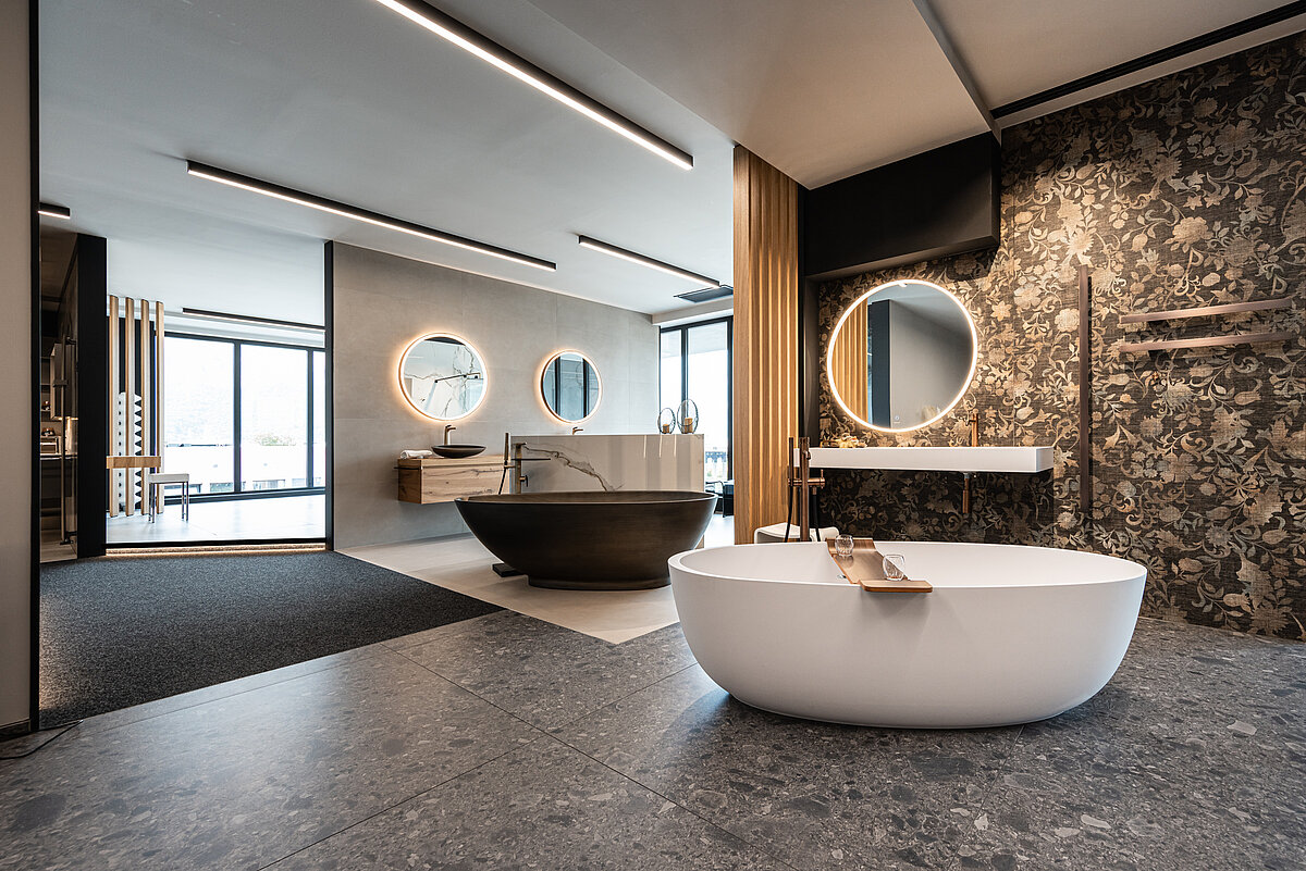 Modernes Badezimmer mit zwei freistehenden Badewannen, einem schwarz-marmorierten und einem weißen. Große runde Spiegel mit Beleuchtung und stilvolle Armaturen. Elegante Fliesen und eine gemusterte Wand ergänzen das luxuriöse Ambiente.