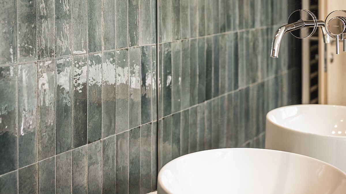 Modernes Badezimmer mit zwei weißen Aufsatzwaschbecken, einer Wand mit glänzenden grünen Fliesen, und eleganten, metallischen Hängeleuchten.