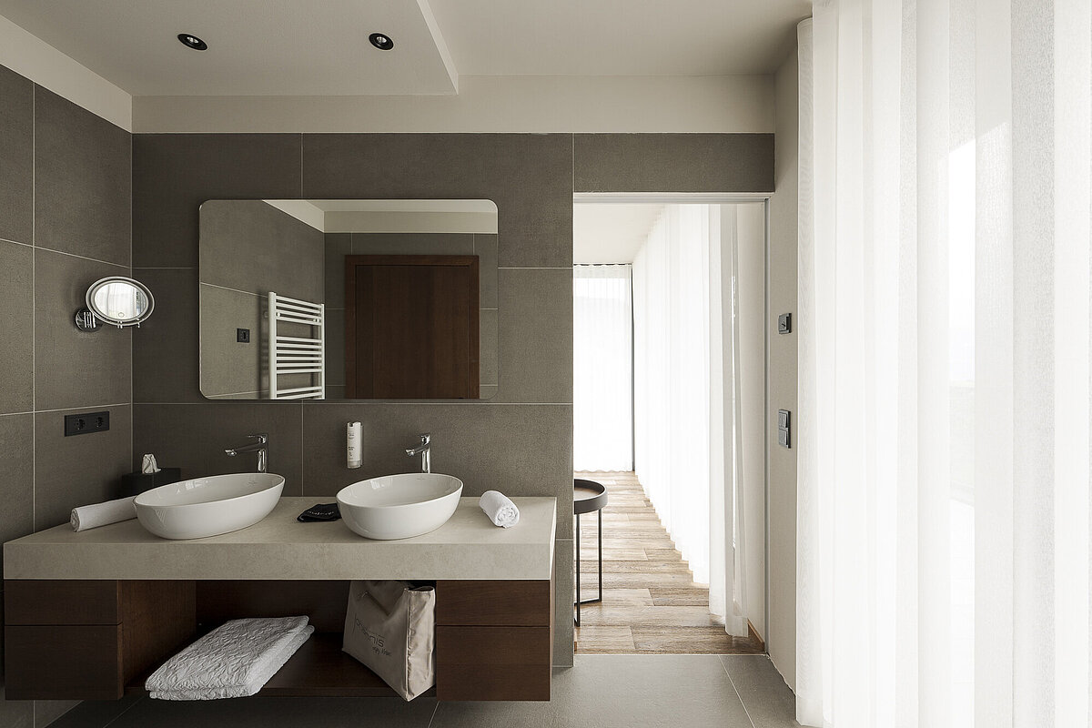 Modernes Badezimmer mit zwei Waschbecken auf einer grauen Arbeitsplatte, einem großen Spiegel an der Wand und einem Handtuch. Neutrale Farbtöne, minimalistisches Design, helle Beleuchtung durch große Fenster mit weißen Vorhängen.