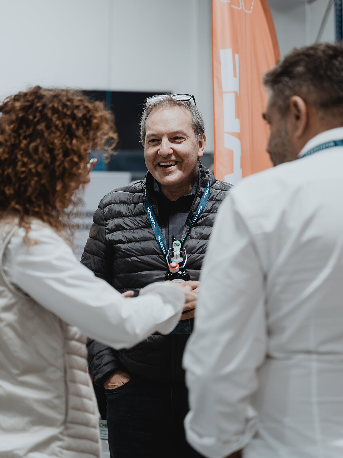 Ein Mann lächelt, während er sich mit zwei anderen Personen unterhält. Er trägt eine schwarze Jacke und hat ein Lanyard um den Hals. Eine Frau mit lockigem Haar steht ihm zugewandt, während eine weitere Person mit kurzem Haar daneben steht.